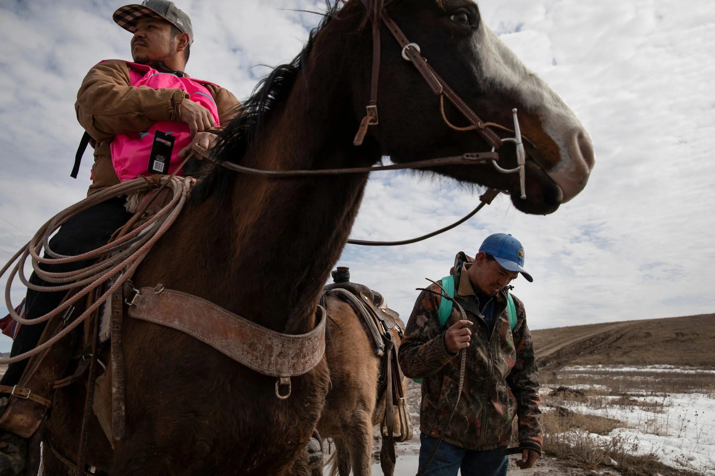 A man and a woman standing outdoors with a brown and white horse. The man is wearing a camouflage cap and jacket, looking down, while the woman is on the horse, wearing a pink backpack and a brown jacket, looking into the distance. The sky is cloudy 