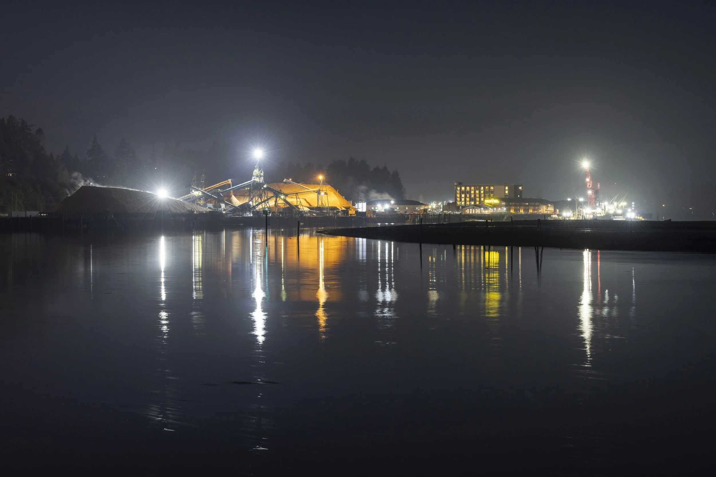 Nighttime view of a harbor with industrial buildings, bright lights, and reflections on the water.