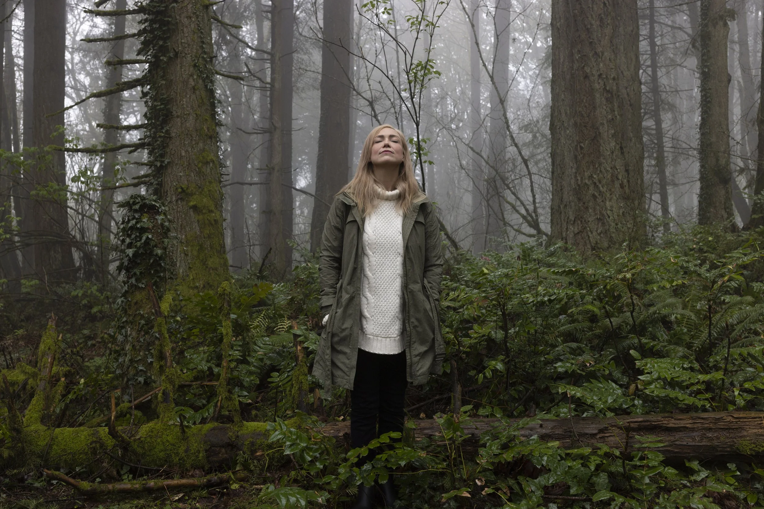 A woman standing in a misty forest with her eyes closed, wearing a green jacket over a white sweater.
