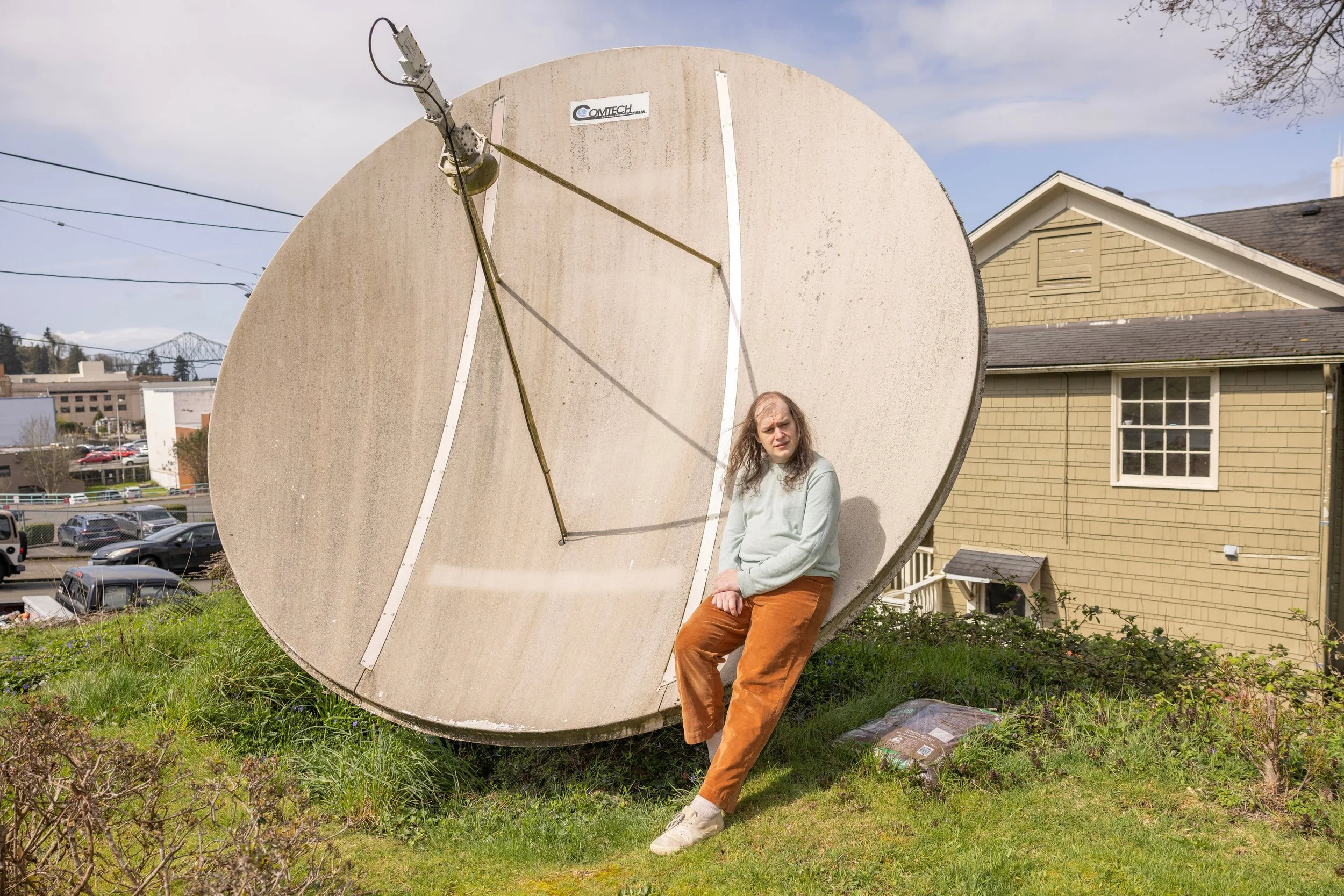 A woman standing and sitting in front of a large satellite dish outside on a grassy area, with houses and trees in the background.