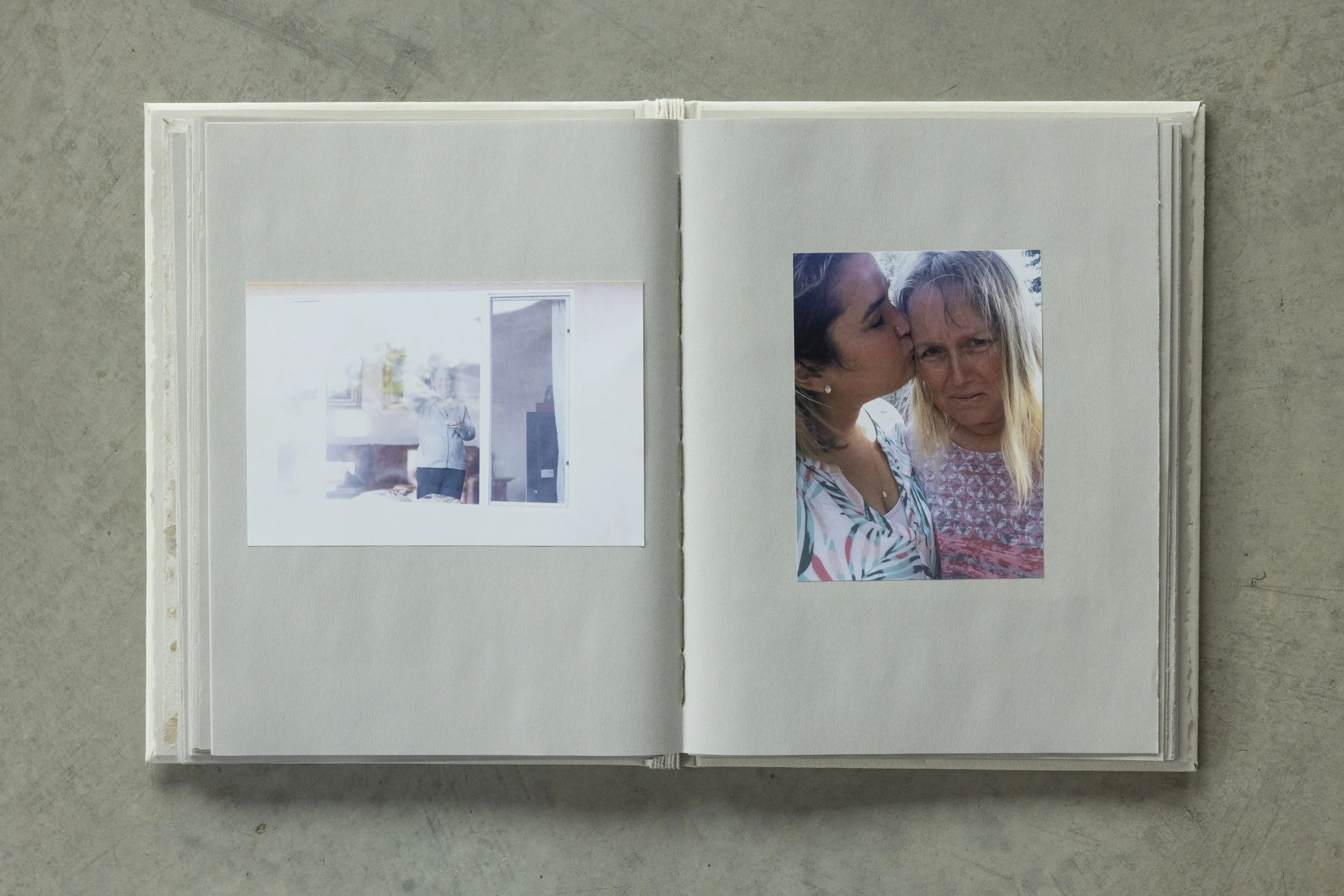 Open photo album with two photographs: the first shows a person standing in a room, the second shows two women, one kissing the other's forehead