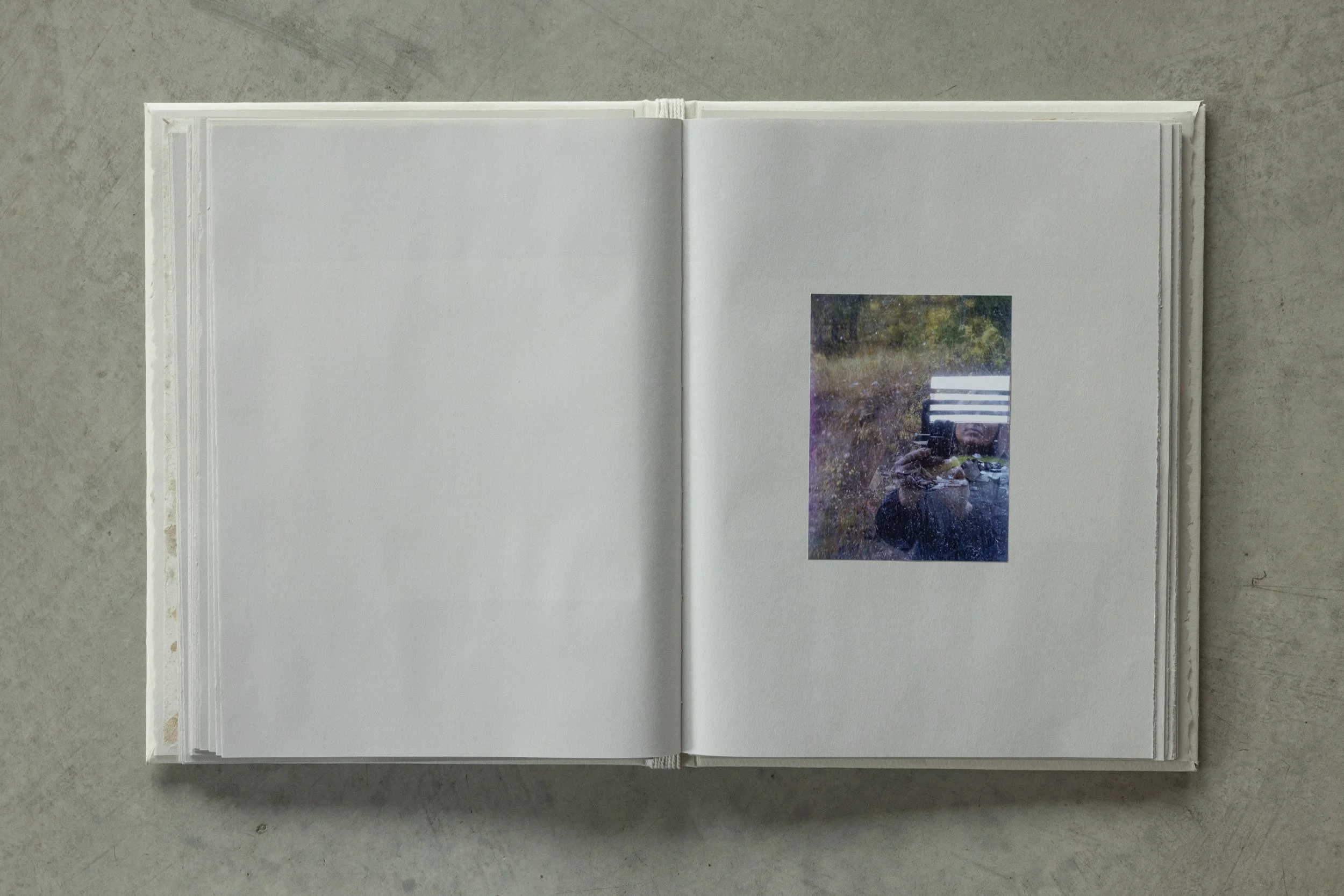 Open white photo album laying on a gray concrete surface, with a small rectangular photograph on the right page showing an outdoor scene with trees and a person taking a picture.