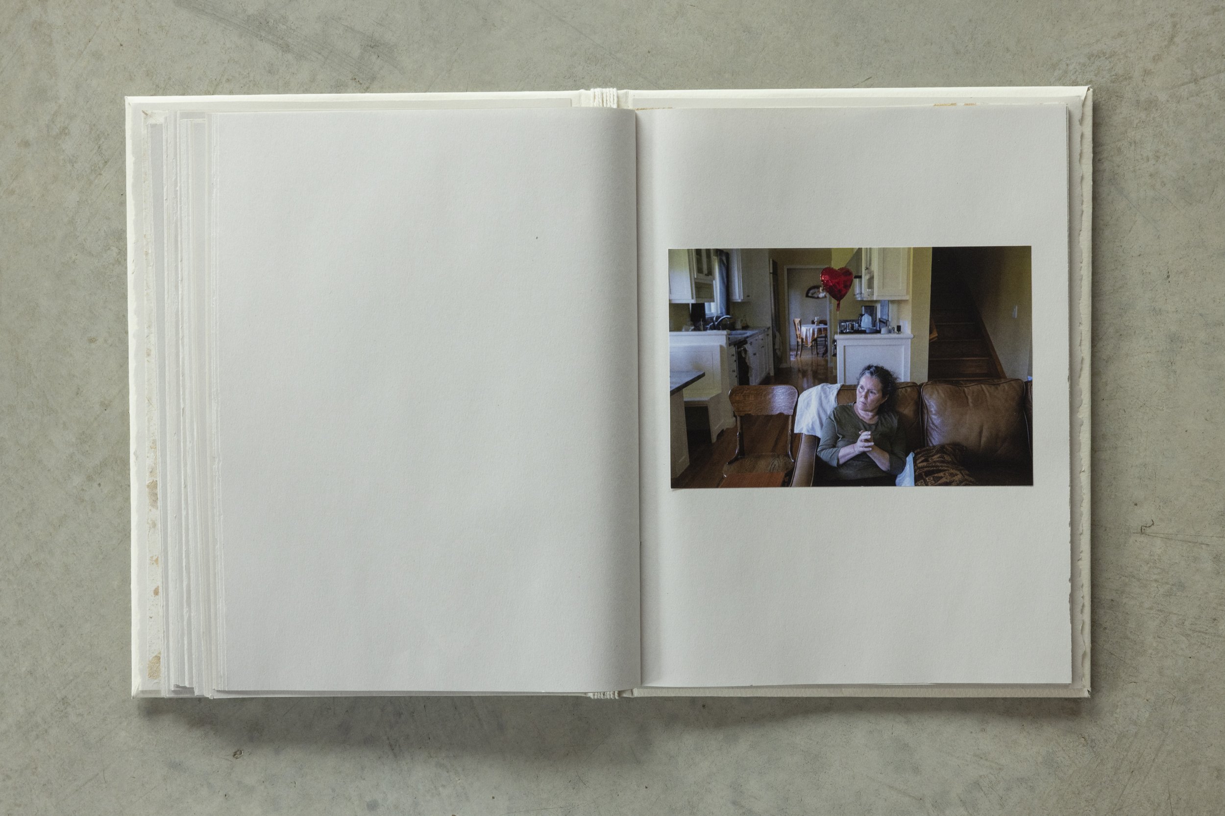 An open photo album with a picture of a woman sitting on a brown leather couch in a cozy kitchen or dining area, holding a cup, with a red heart-shaped balloon floating nearby.
