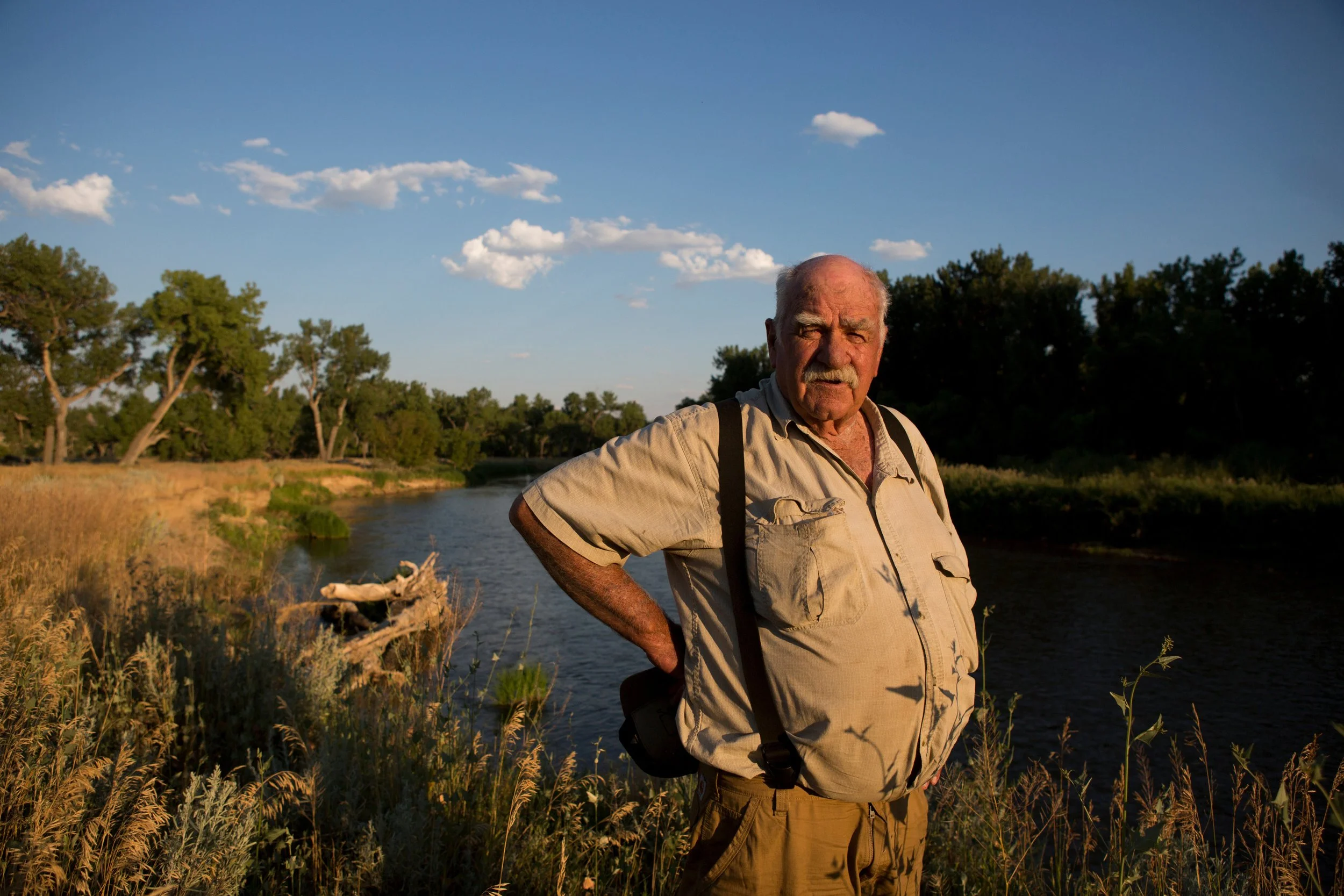 An elderly man standing outdoors near a river, with trees and a partly cloudy sky in the background, dressed in a beige shirt and khaki pants.