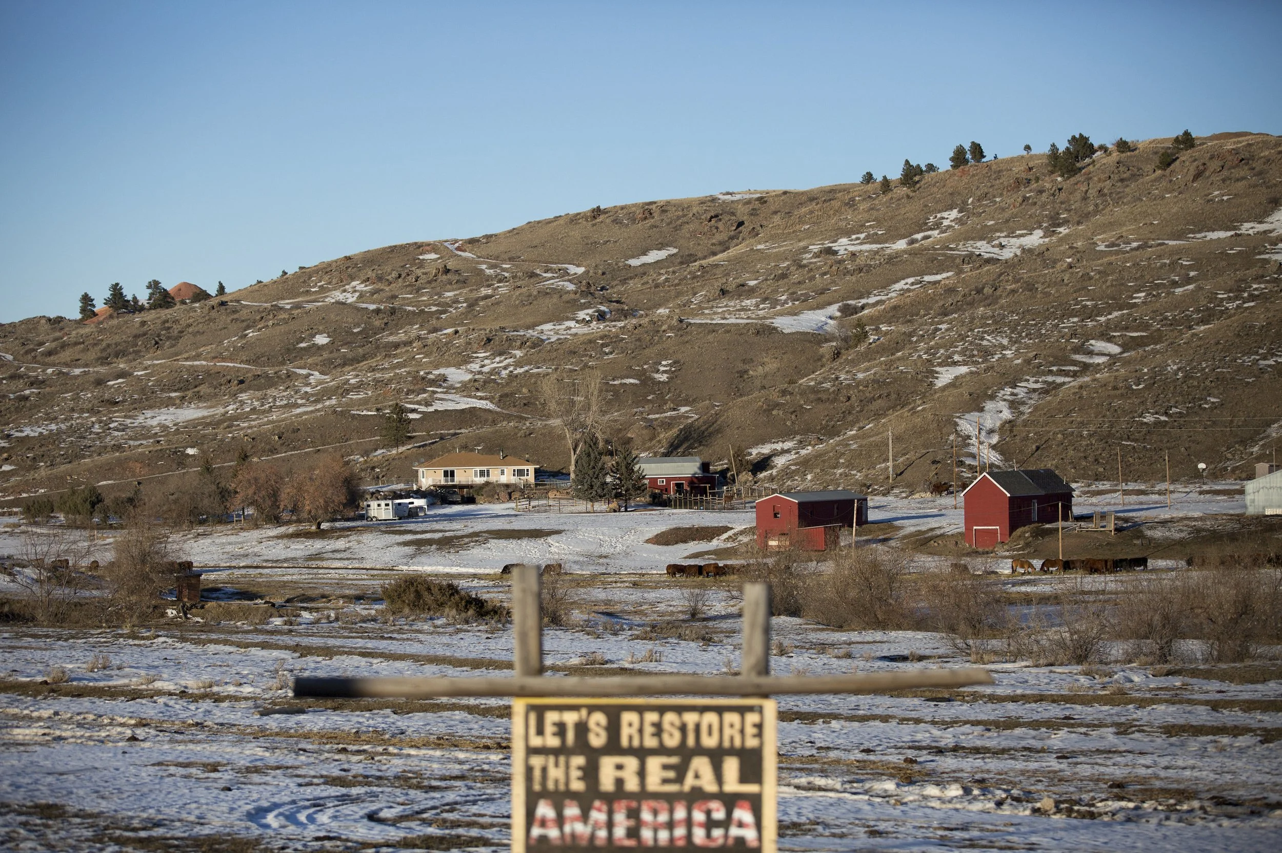 A rural landscape with snow patches on hills, some trees, farm buildings including a red barn, and horses on the field. There is a sign in the foreground that reads 'Let's restore the real America'.