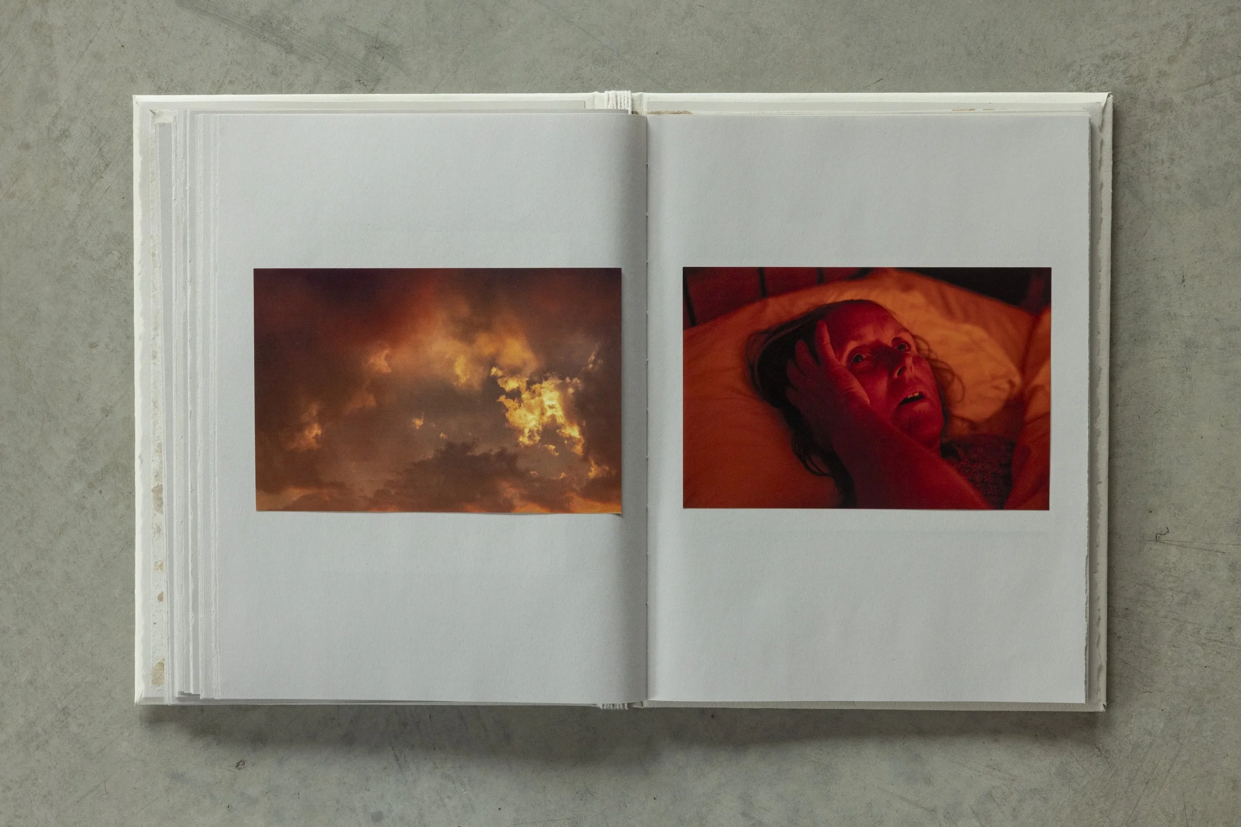 Open photo book with two pages visible. The left page has an image of a cloudy sky during sunset or sunrise with orange and yellow clouds. The right page shows a woman lying in bed, touching her head, with a red-lit background.