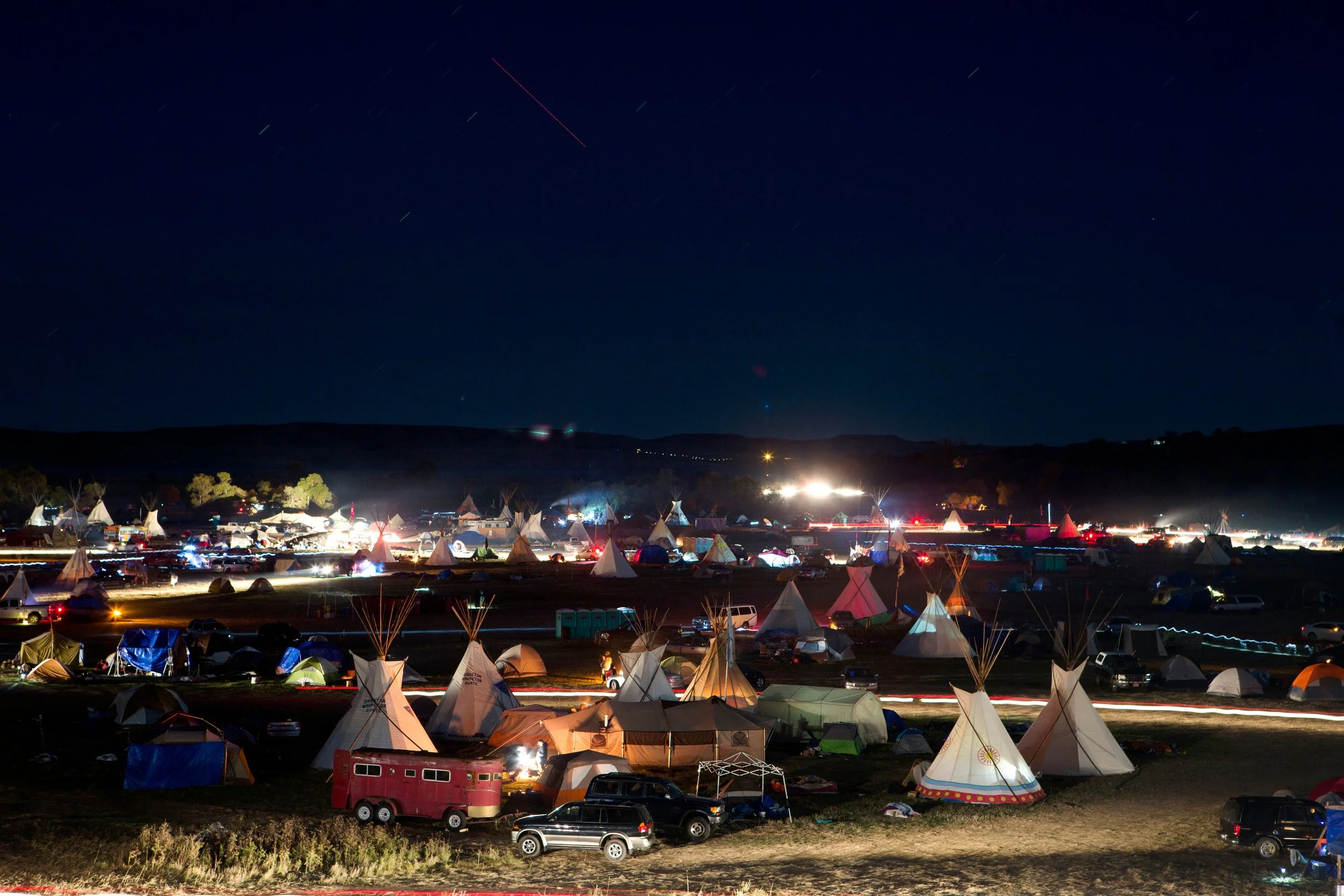 Nighttime scene of a large outdoor camping event with numerous tents, vehicles, and trailers illuminated by artificial lights, set against a dark sky with stars at the Dakota Access Pipeline water protectors' camp. 