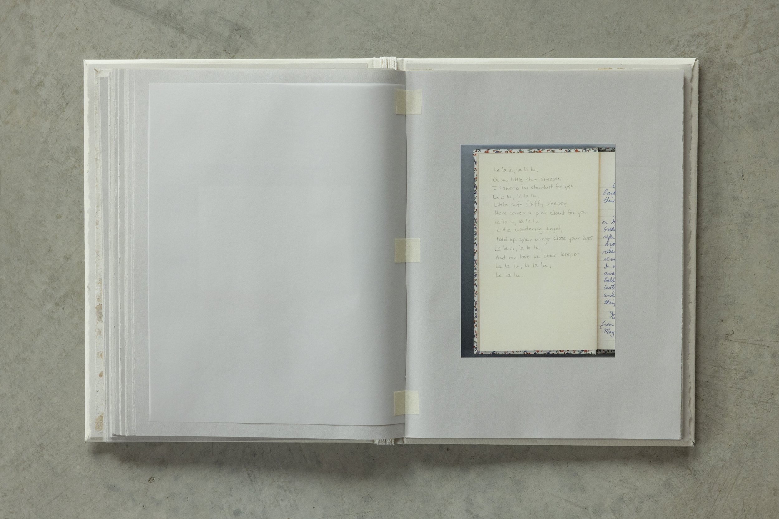 An open notebook with tape inside, laying on a concrete surface. The right page shows a small, decorated photo or card with handwritten lyrics to a lullaby.