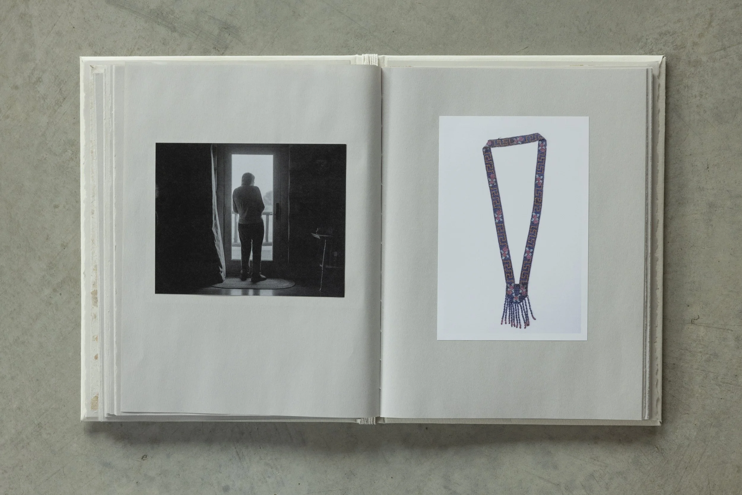 Open book with a black-and-white photo of a person looking out a window on the left page and a color photograph of a beaded necklace on the right page, on a concrete surface.