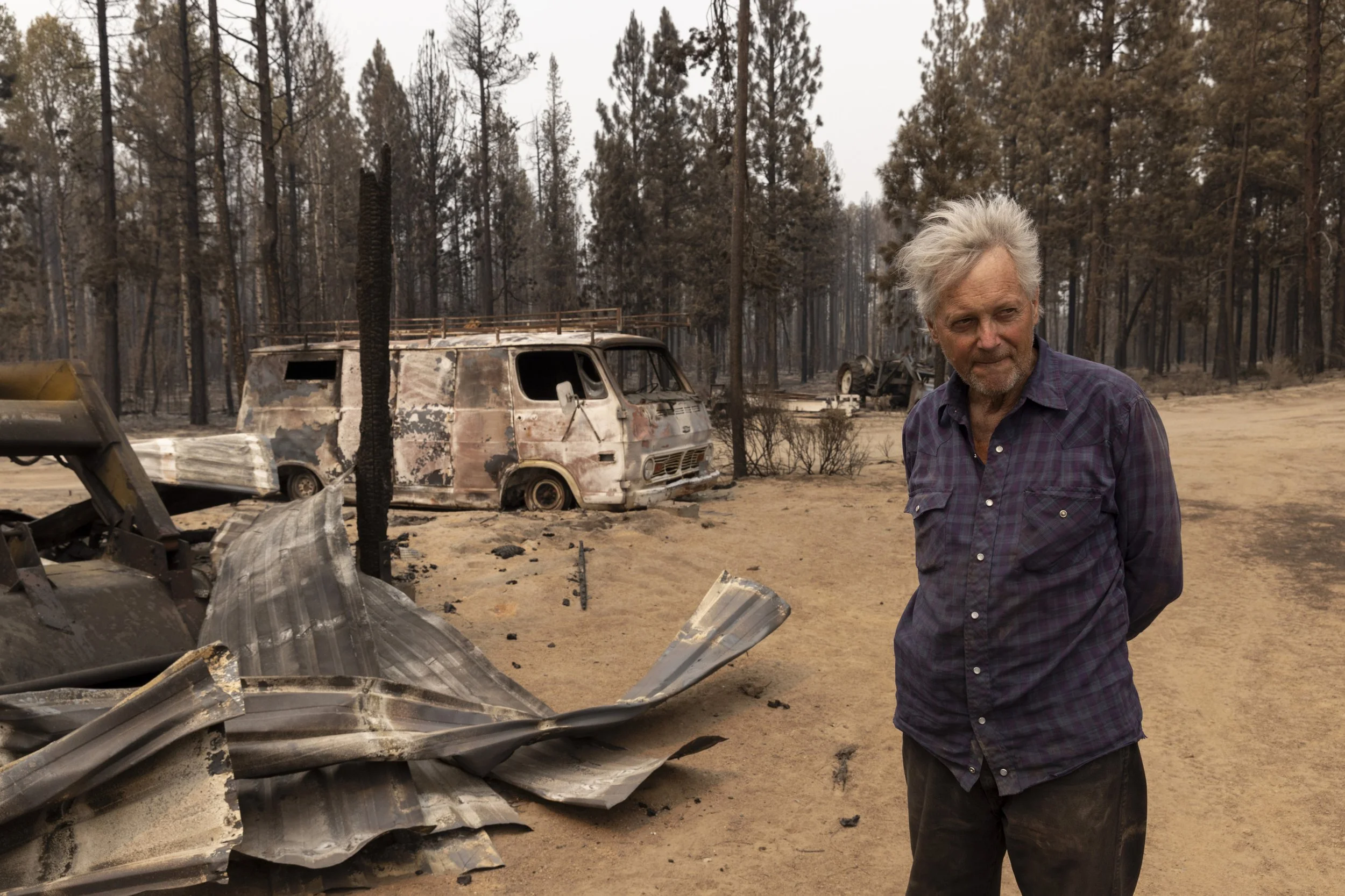 A man with gray hair and a plaid shirt stands in front of burned trees and wreckage of a burnt vehicle in a forest after a wildfire.