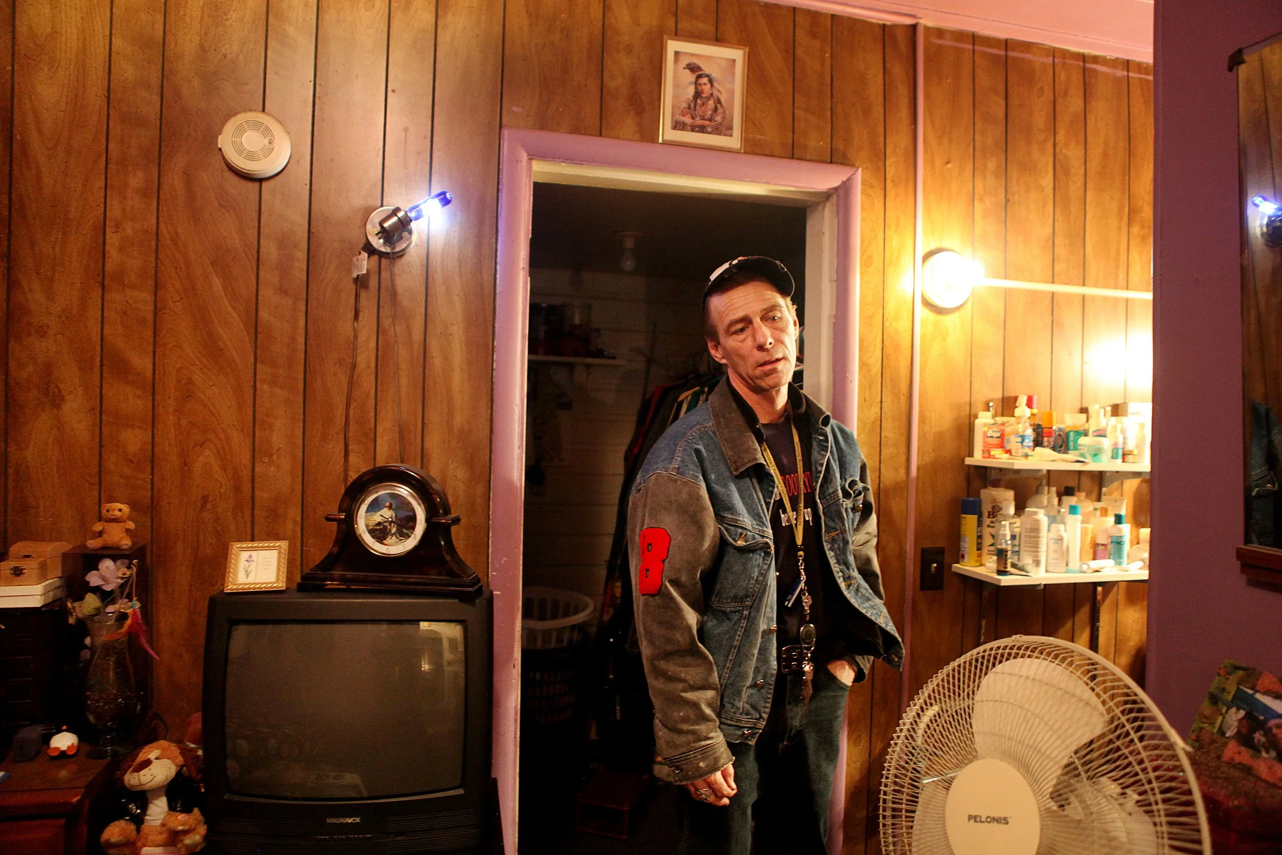 A man wearing a denim jacket and a hat stands inside a room with wood-paneled walls, looking at the camera. Behind him, there is a purple doorway, a shelf with bottles, a small framed artwork, and a vintage TV set. Various decorations, including tedd
