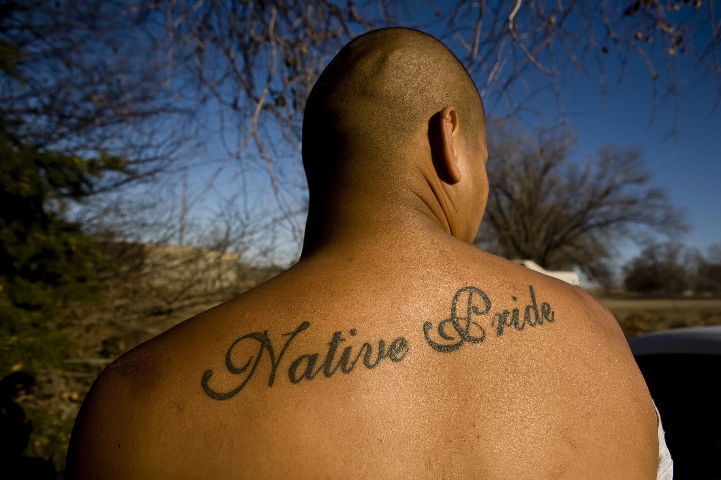 Close-up of a person's back and shoulder, with a tattoo that reads 'Native Pride' in cursive, taken outdoors during the day with a background of leafless trees and blue sky in North Dakota.