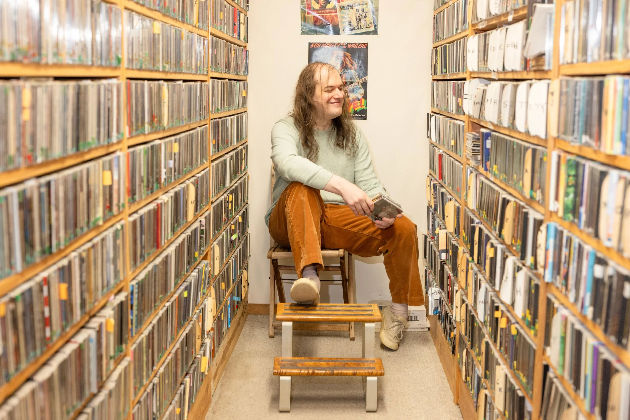 A woman with long hair and a smile, sitting on a wooden bench in a narrow library aisle filled with shelves of music CDs. She is holding a book and has a cheerful expression. 