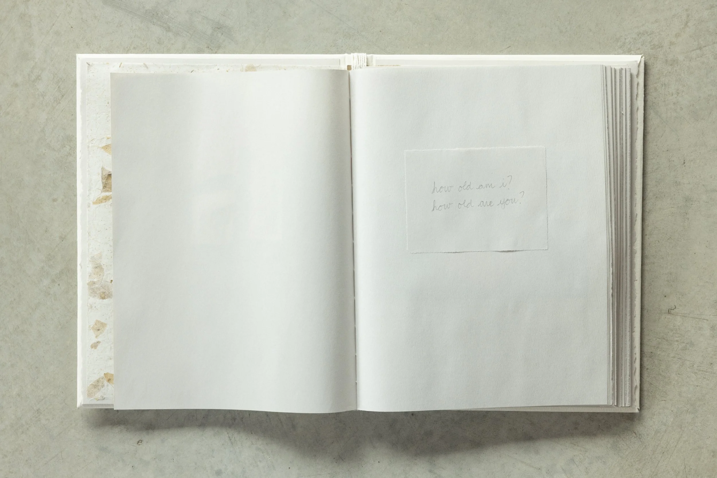 Open notebook with blank pages on a textured surface, with the right page containing faint handwritten question: 'how old am I? how old are you?'