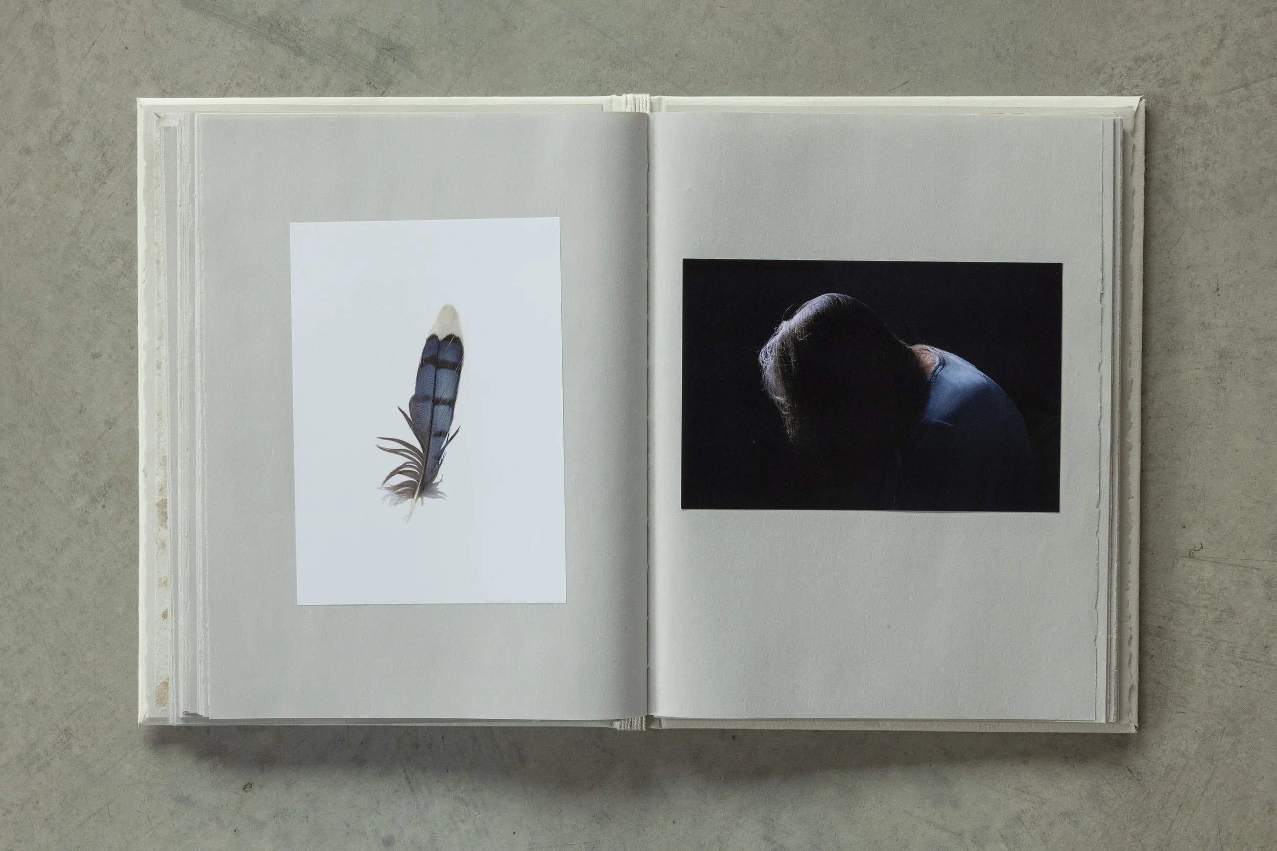 Open book featuring a photograph of a person with long hair in a dark shirt on a black background and a print of a feathered wing on the opposite page, placed on a gray surface.