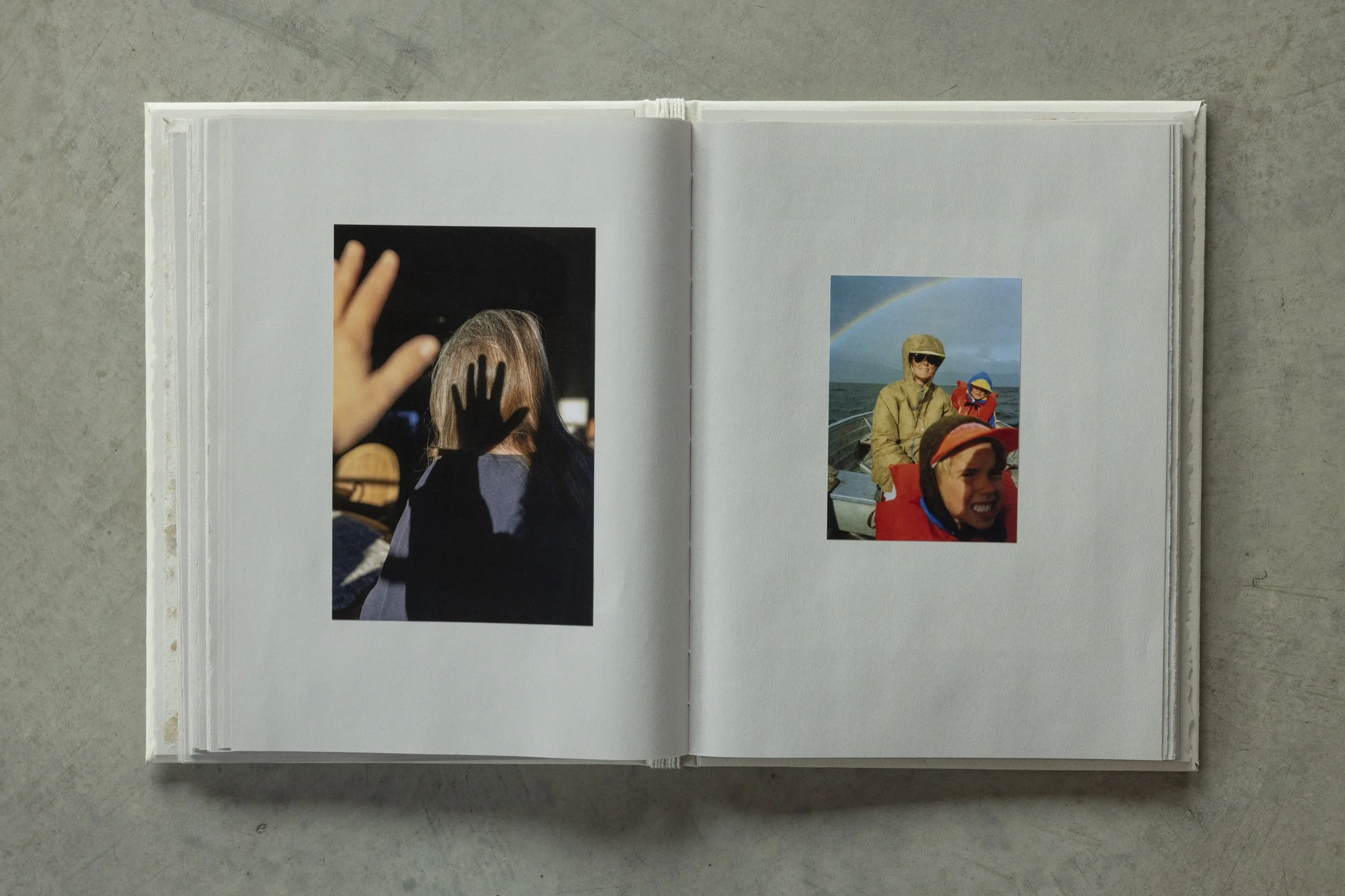 Open scrapbook with two photographs on a gray surface. The left photo shows a person with gray hair and a hand raised, and the right photo depicts people on a boat with a rainbow in the background.