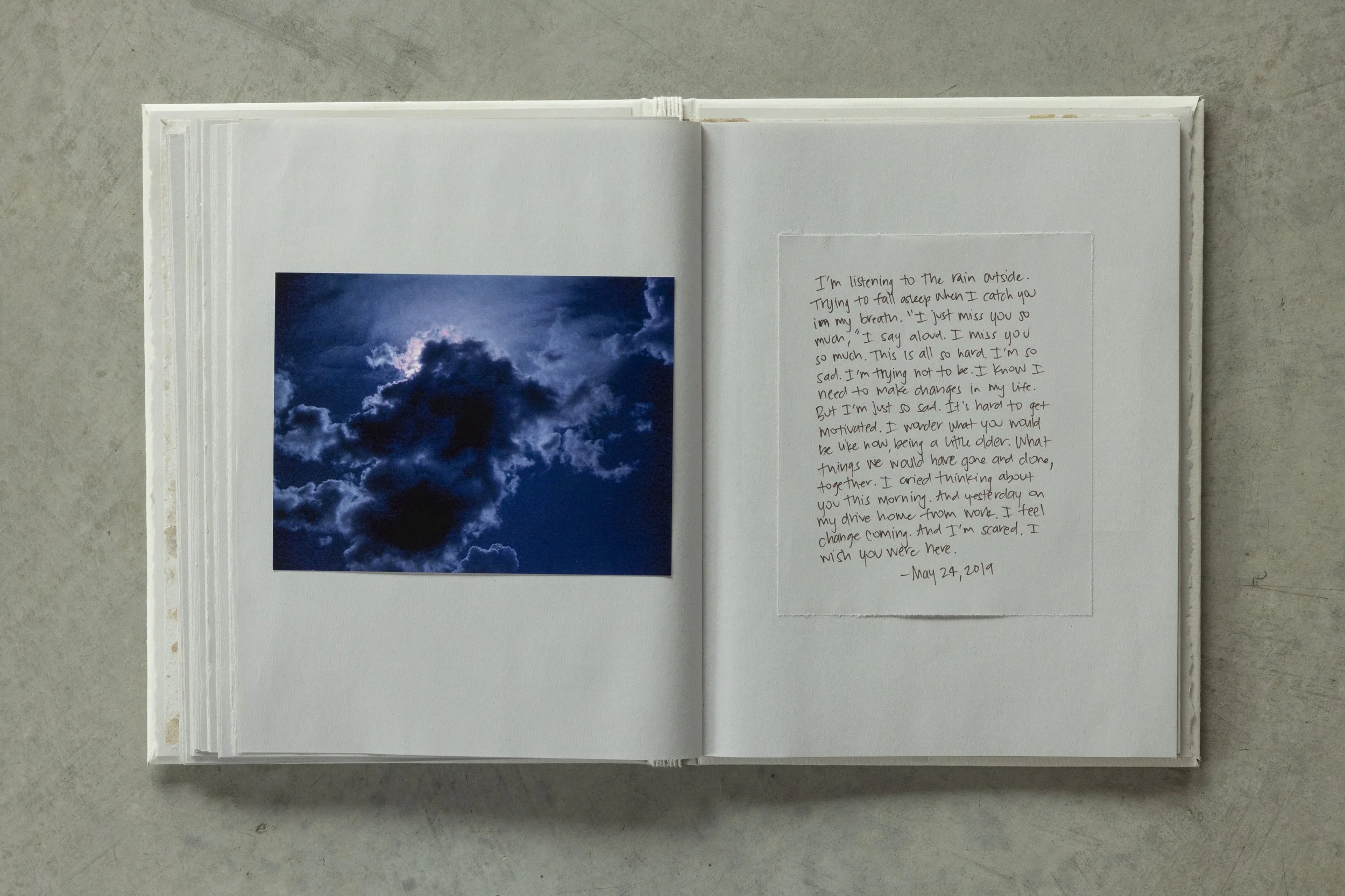 Open white scrapbook or journal on a plain surface, with a photograph of dark, cloudy sky on the left page and handwritten text on the right page.