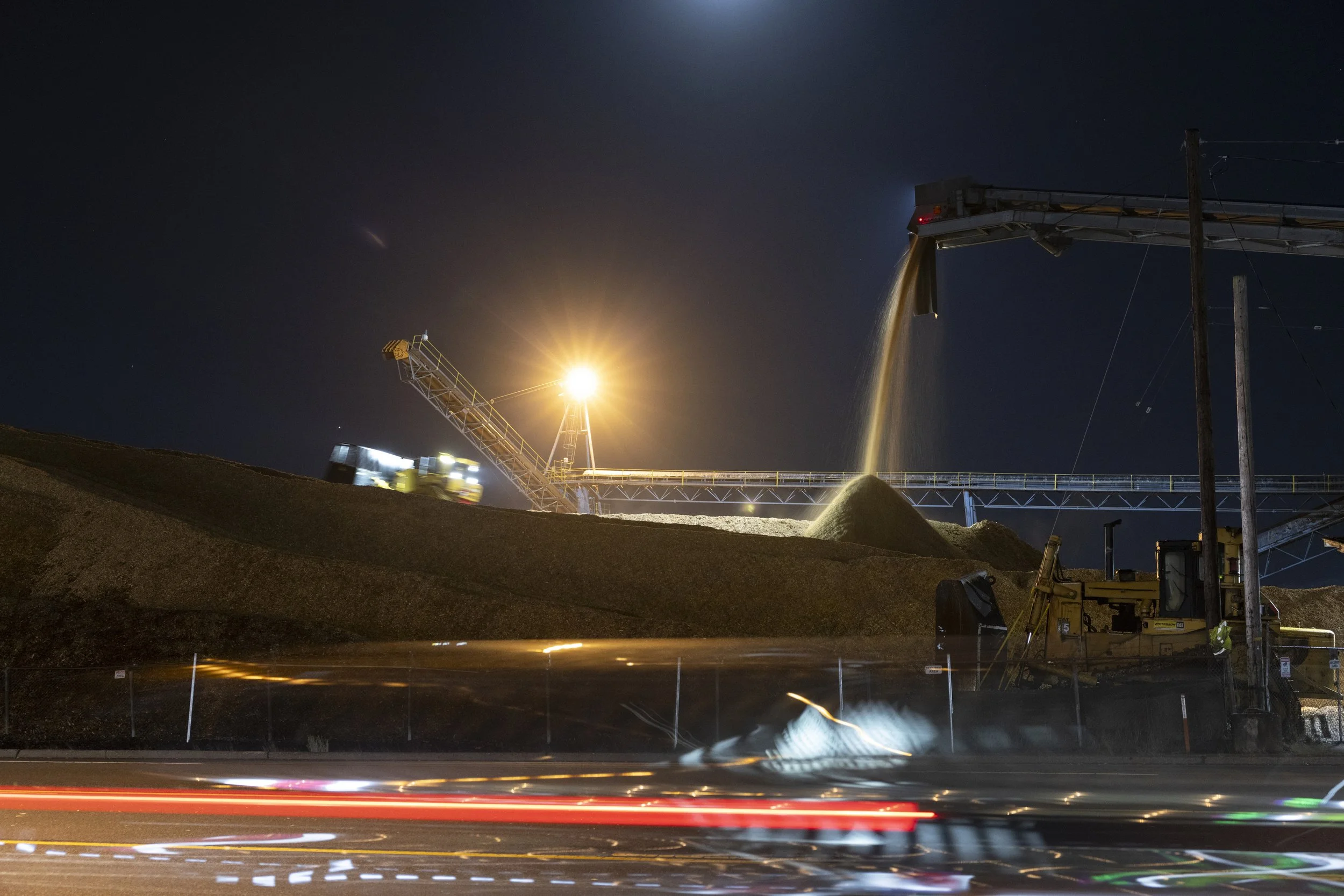 Nighttime scene of an industrial site with a conveyor system pouring granular material onto a pile, illuminated by bright lights, with blurred vehicle light trails in the foreground.