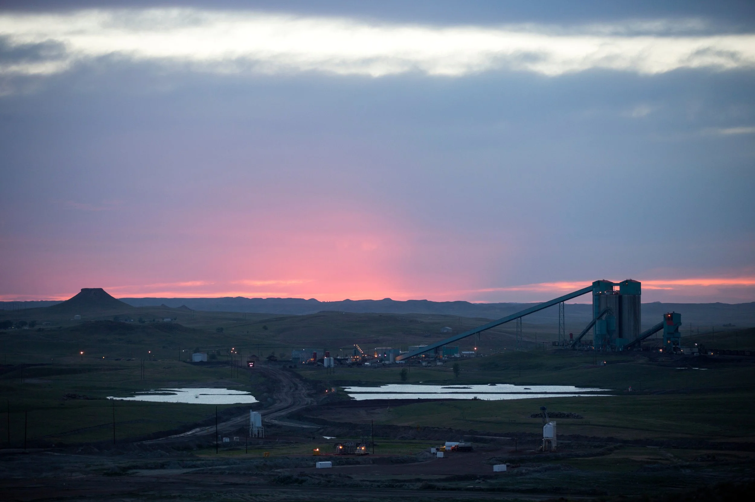 A landscape view of a rural area at dawn with a factory in the foreground, some bodies of water, and a mountain in the distance under a sky with clouds and a pink hue from the sunset.