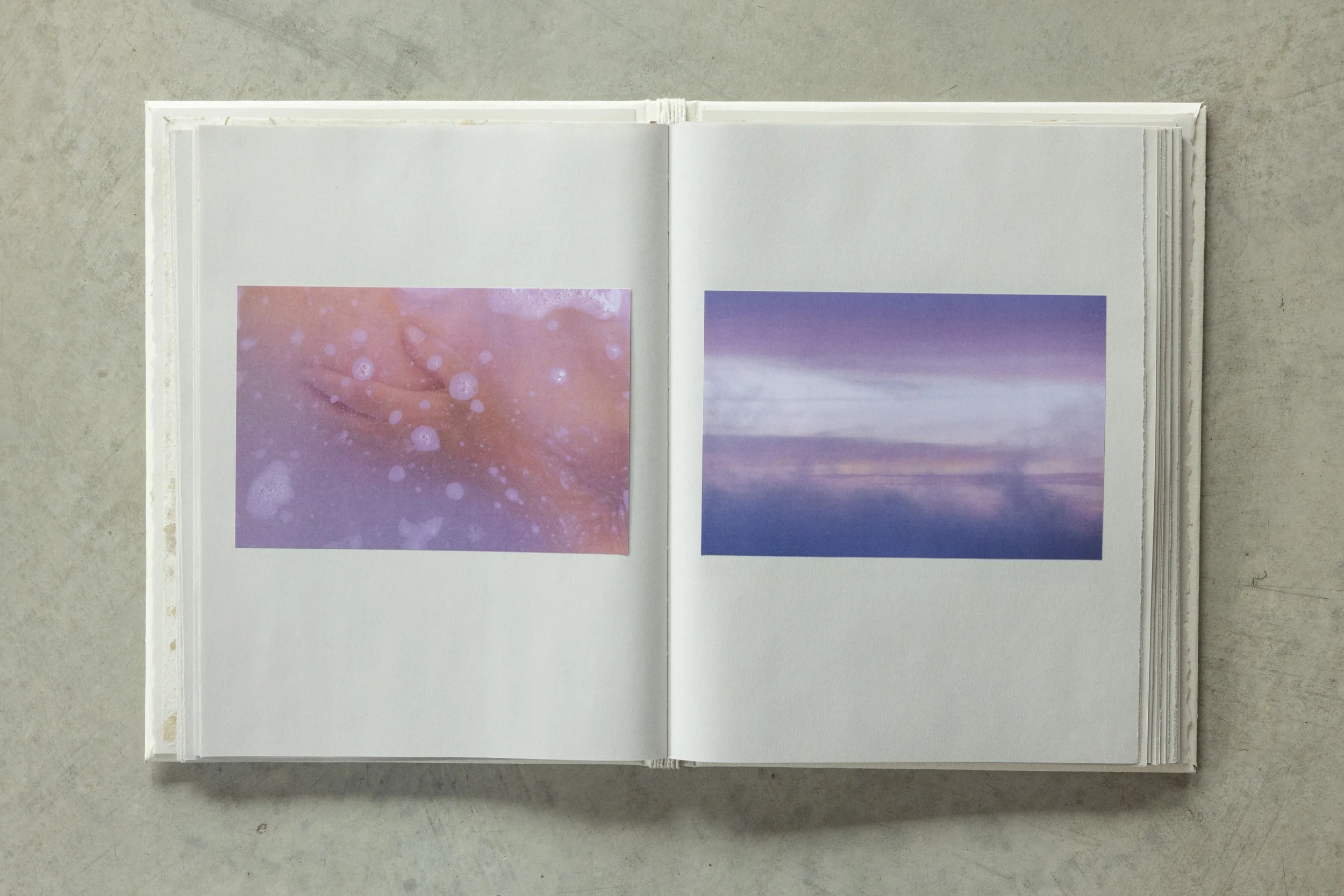 An open white photo album with two photographs on plain white pages, placed on a gray surface. The left photo shows an out-of-focus pinkish scene with bubbles, and the right photo depicts a purple sky with clouds.