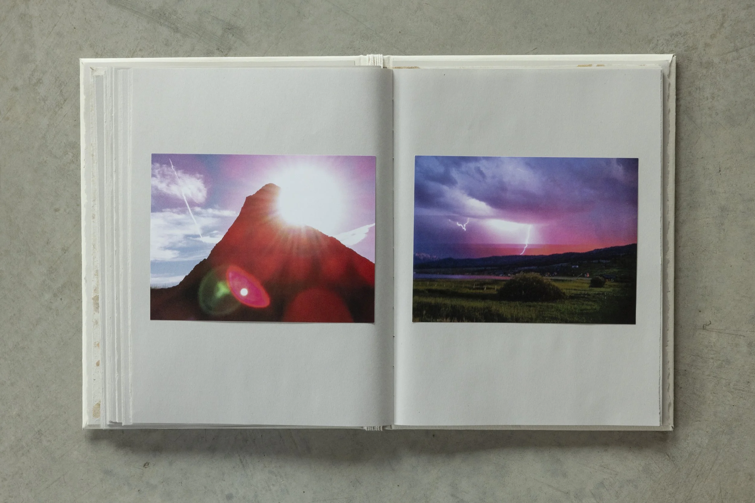 Open photo book displaying two landscape photographs, with mountain and lightning scenes, on a concrete surface.