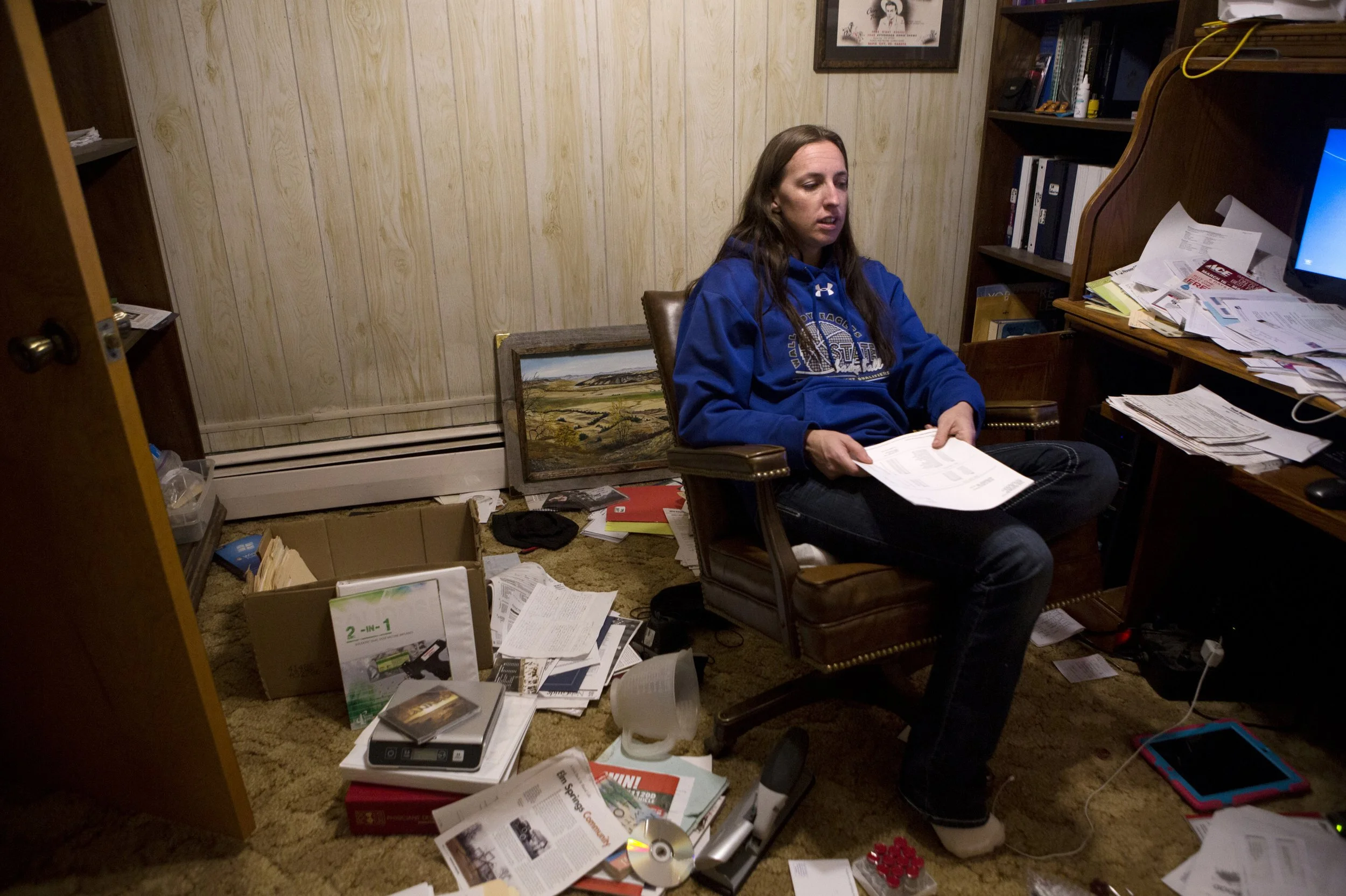 A woman in a blue hoodie sitting in a cluttered room surrounded by scattered papers, books, and electronic devices, holding a piece of paper.