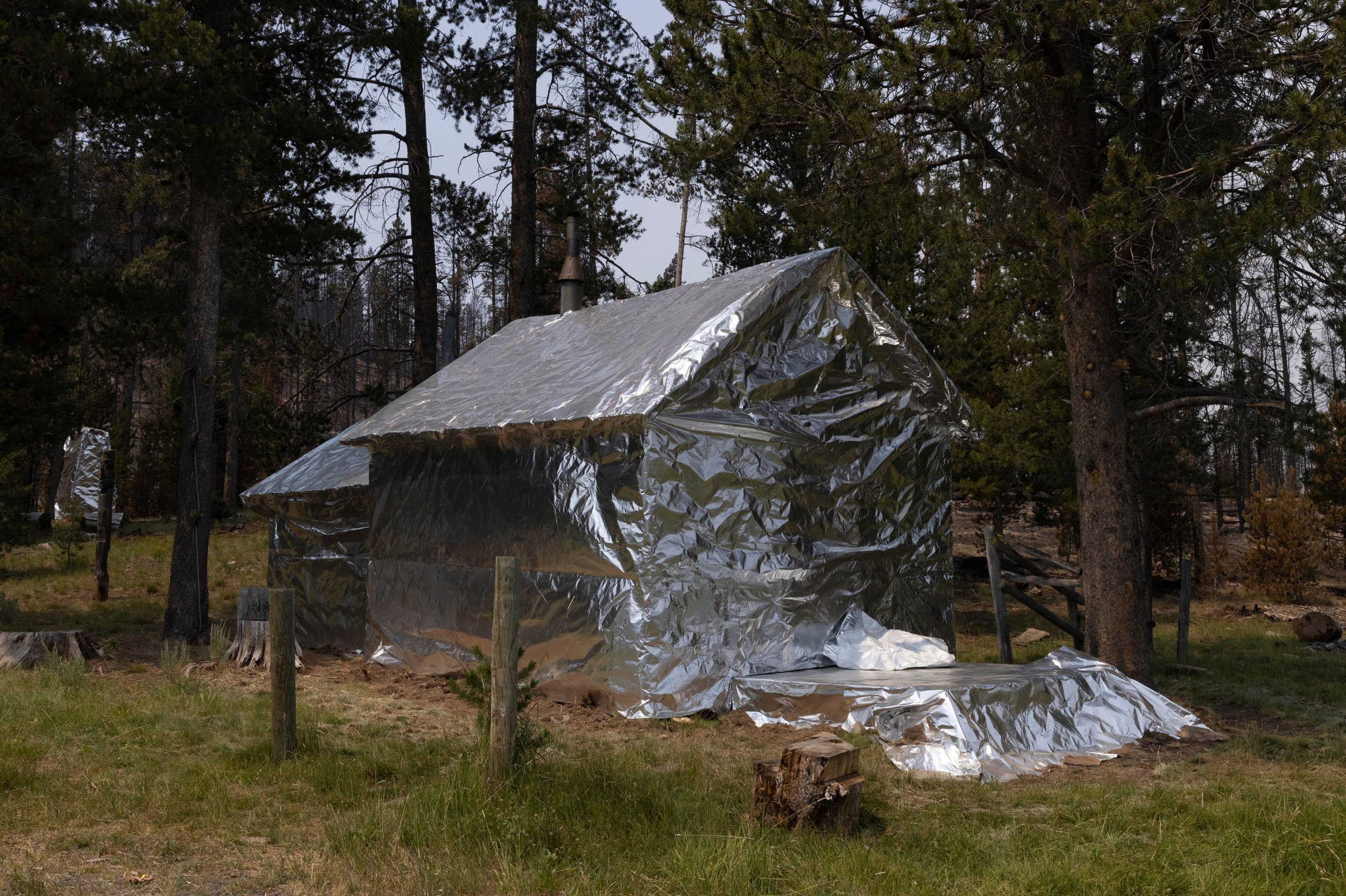 An outdoor scene showing a small house covered in reflective silver insulation material, surrounded by trees, with a chimney on the roof, during daylight, near where wildfires are active in Oregon.