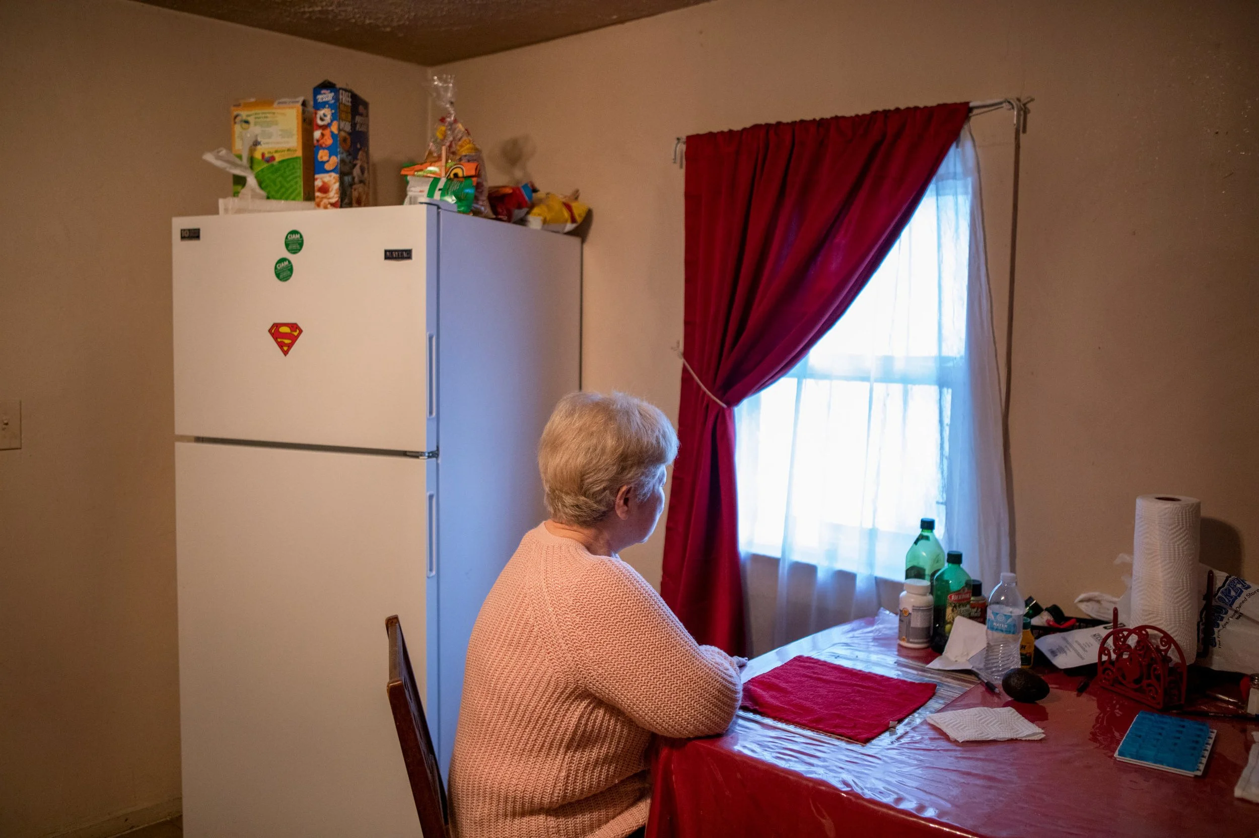 An elderly woman with short white hair sitting at a dining table, facing a window with red and white curtains, surrounded by various bottles and household items.