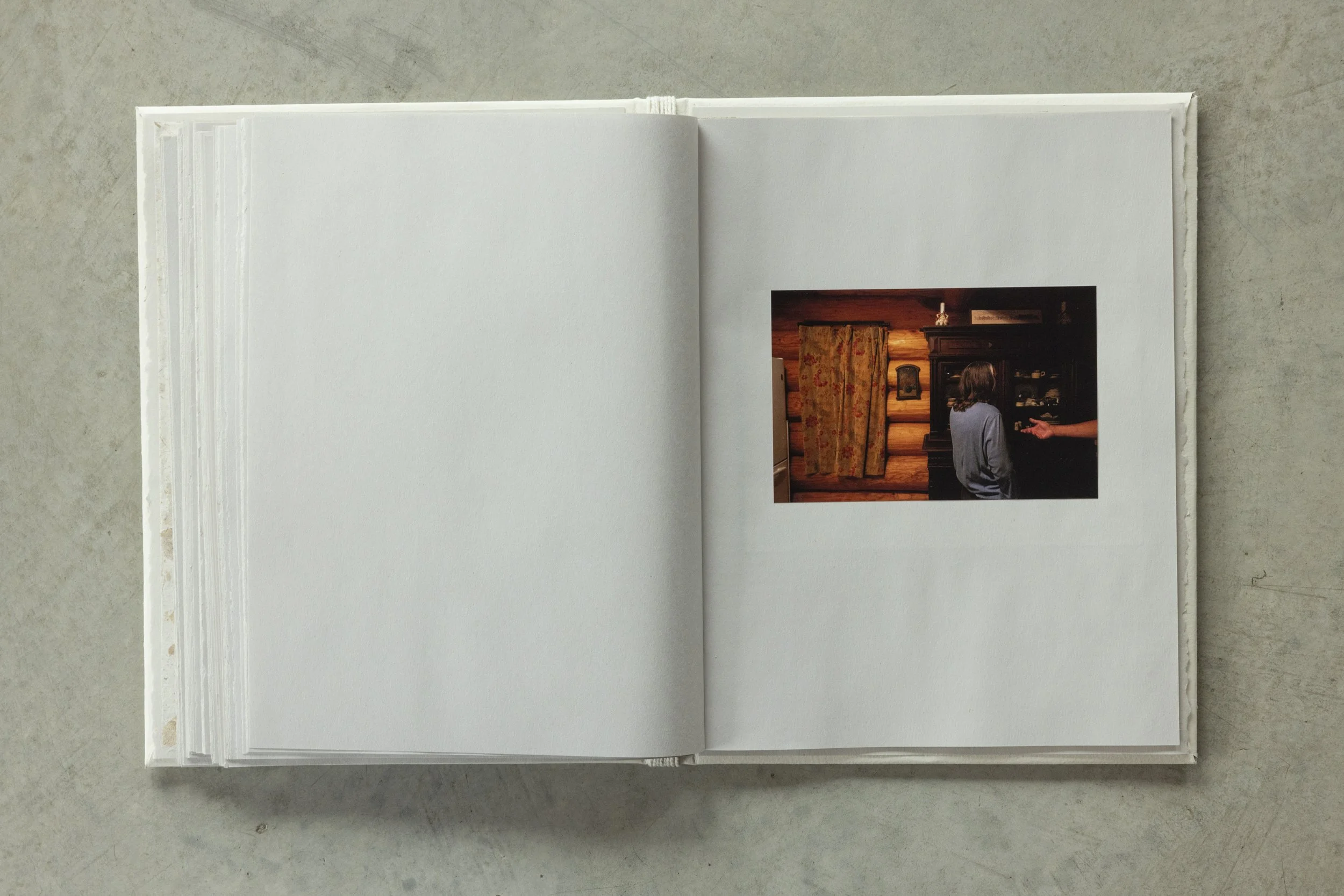Open photo album showing a picture of two people in a cozy, wooden interior, one sitting and one standing, with a dark fireplace and curtains in the background.