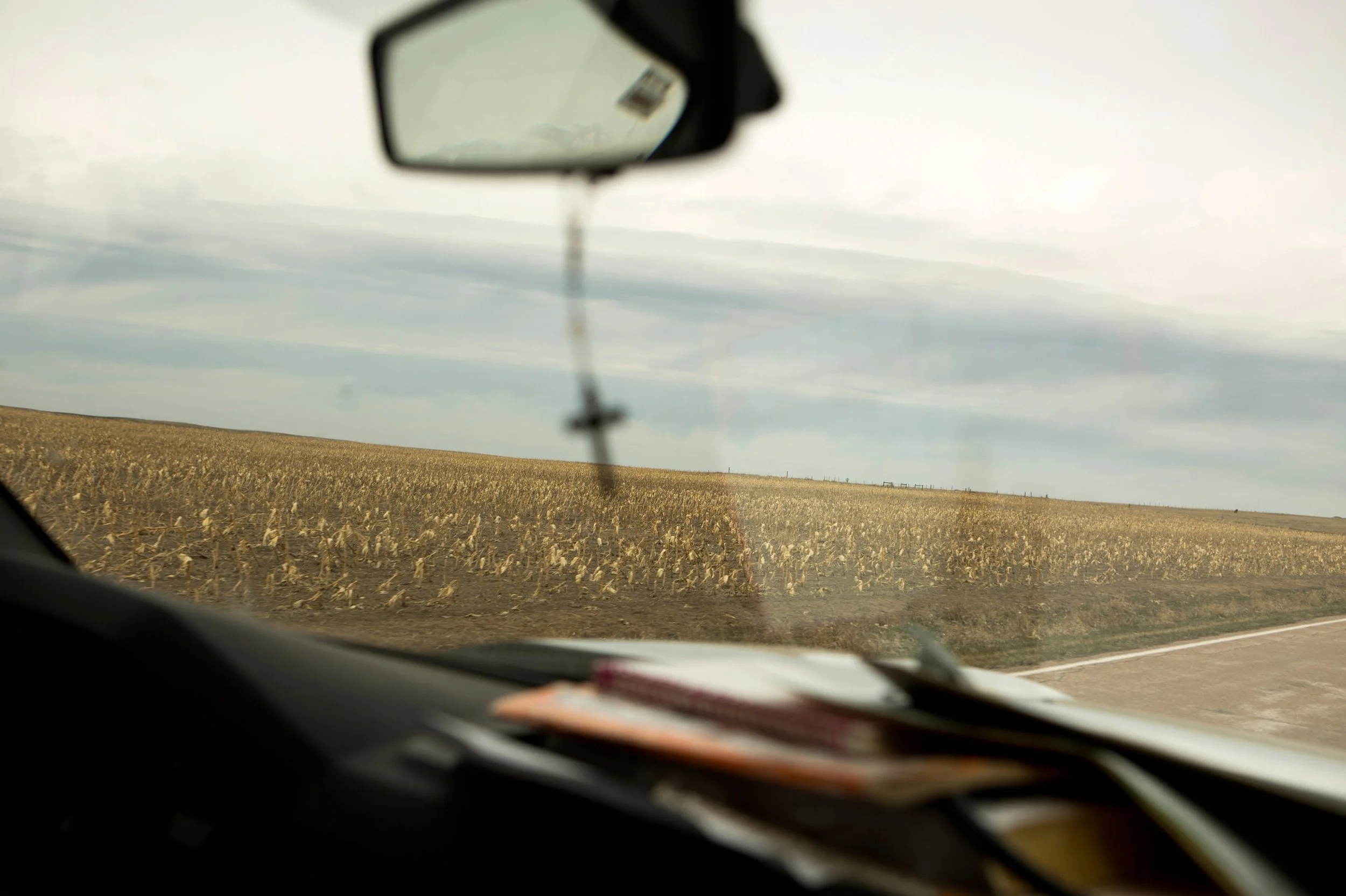 View from inside a vehicle showing a dry, harvested farmland landscape through the front windshield, with a side mirror reflecting a partial view of the sky.