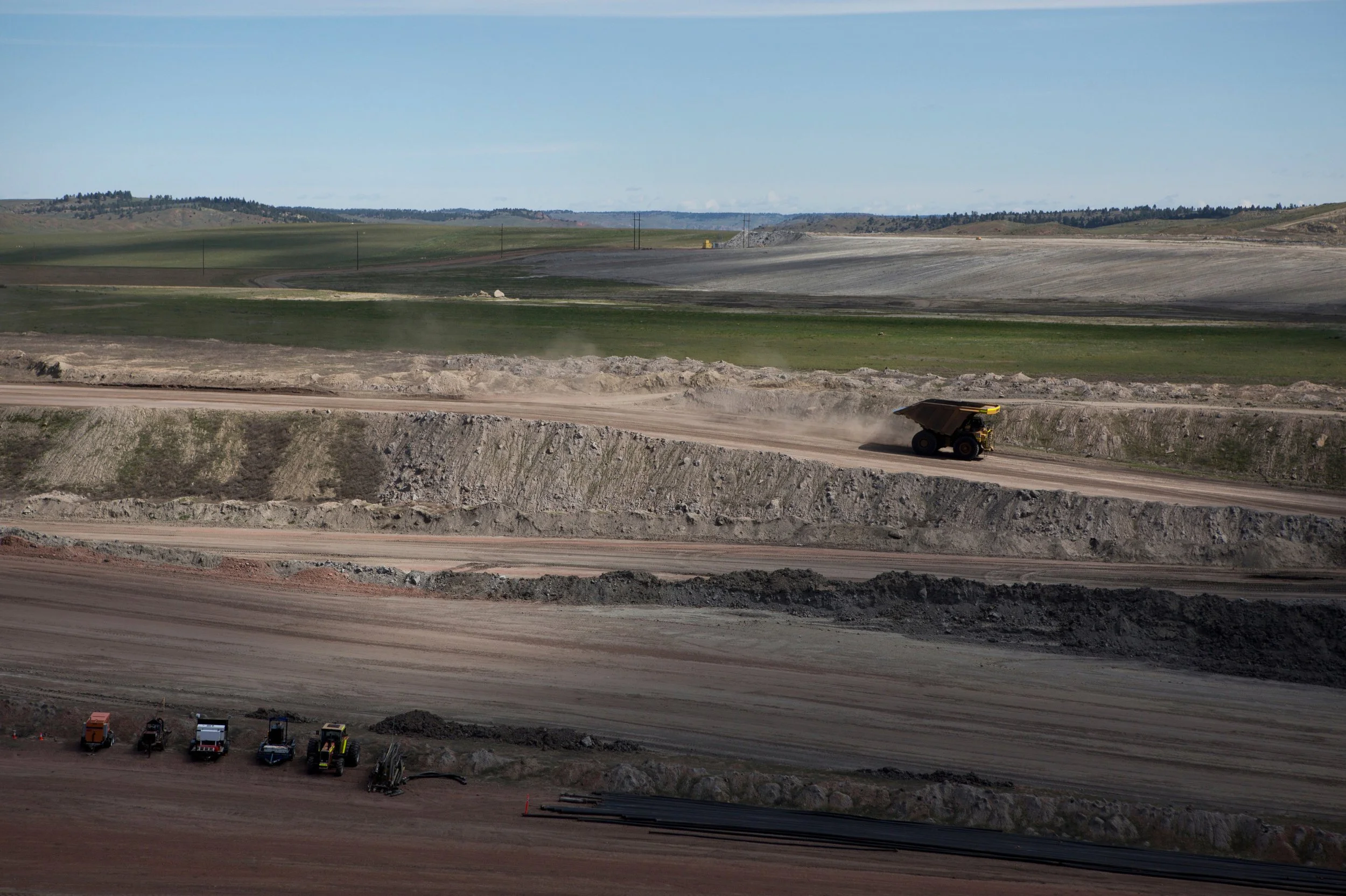 Coal mining site with dirt roads, construction vehicles, and equipment in Wyoming. Large dump truck moving dirt across the site with rolling green hills in the background.
