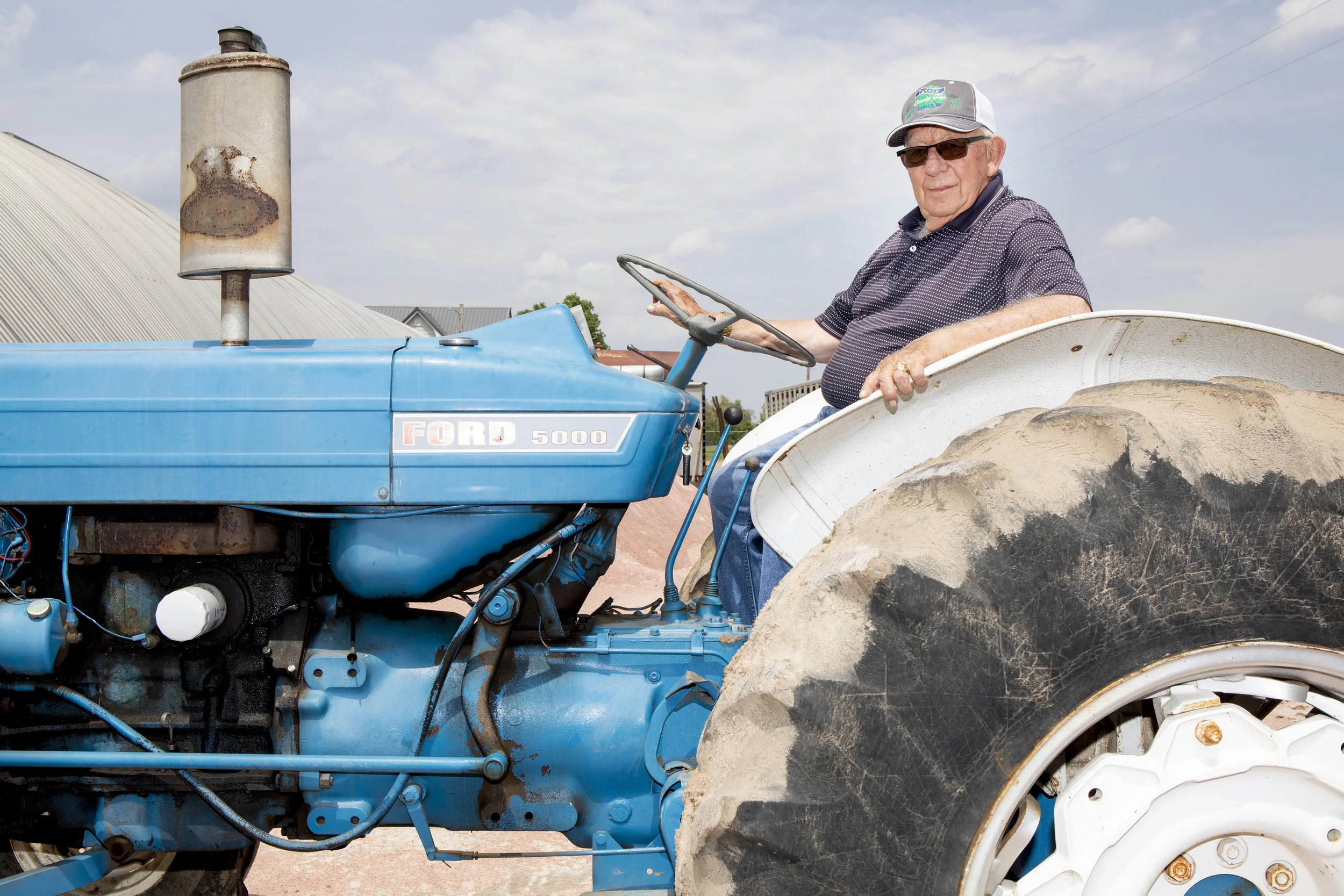An elderly man wearing sunglasses and a baseball cap driving a blue Ford 5000 tractor with large, dusty rear tires on a farm with a cloudy sky.