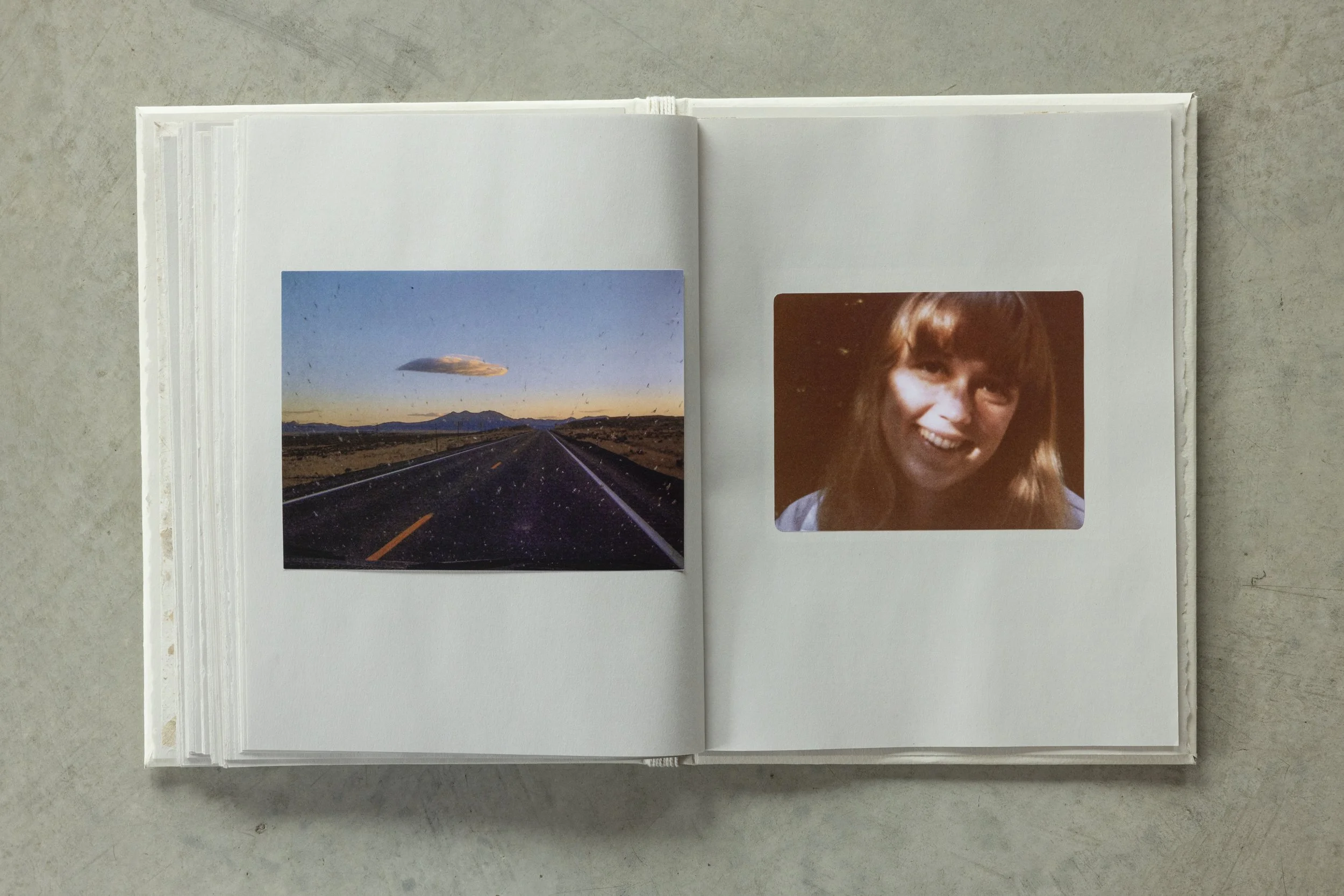 Inside a photo album, on the left page there is a picture of a road stretching into the horizon with mountains in the background and a cloud formation above. On the right page, there is a portrait of a smiling woman with shoulder-length hair.