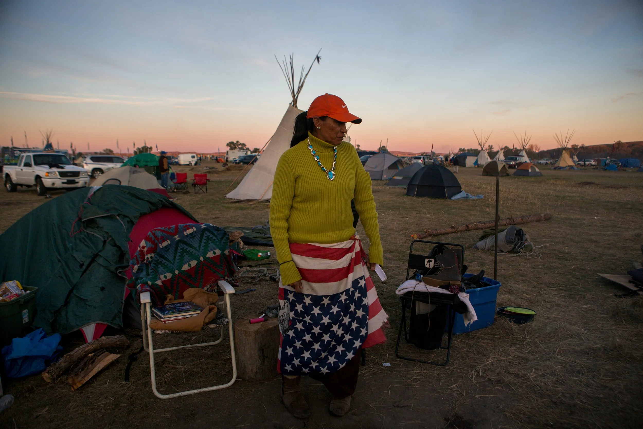 A woman wearing a red cap, yellow sweater, and a flag wrapped around her stands in a campsite at dusk, surrounded by tents and cars at the Dakota Access Pipeline water protectors' camp. 