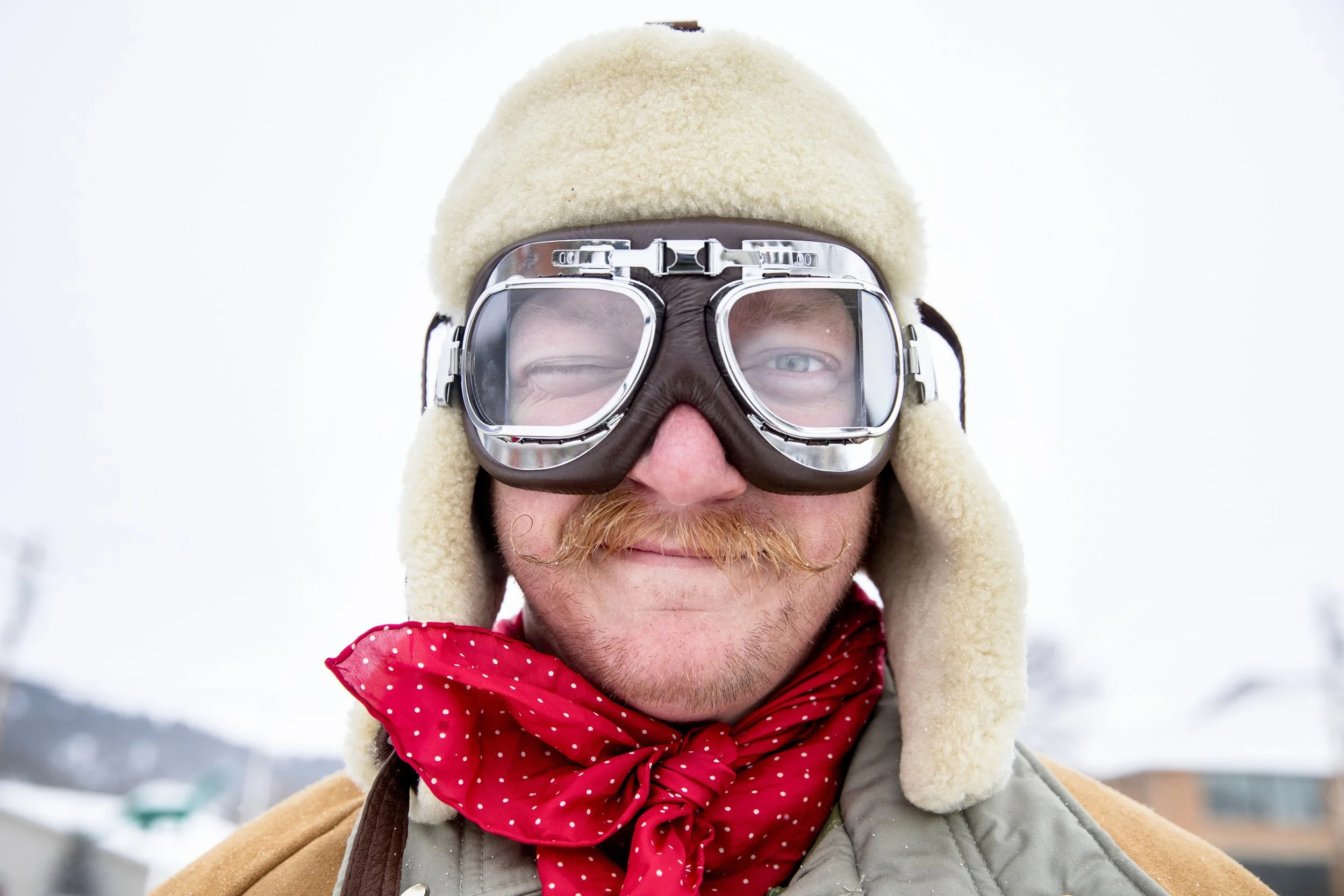 A man with a mustache and beard wearing a beige winter hat with ear flaps, goggles, a red polka dot scarf, and a gray winter jacket outdoors in a snowy environment.