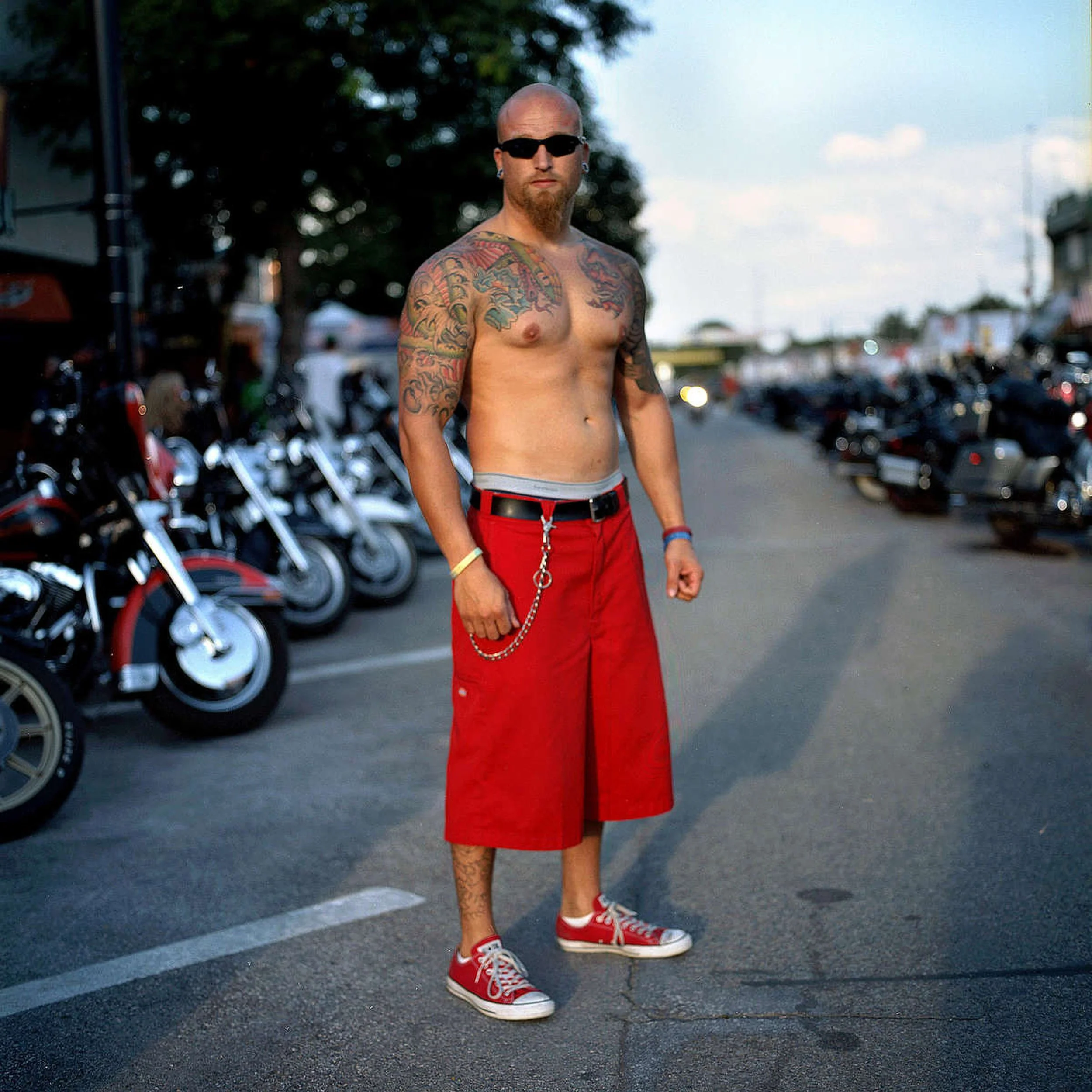 A shirtless man with tattoos standing on a street with parked motorcycles beside him, wearing red shorts, red sneakers, and sunglasses, with a chain hanging from his shorts during the Sturgis Motorcycle Rally in Sturgis, South Dakota.