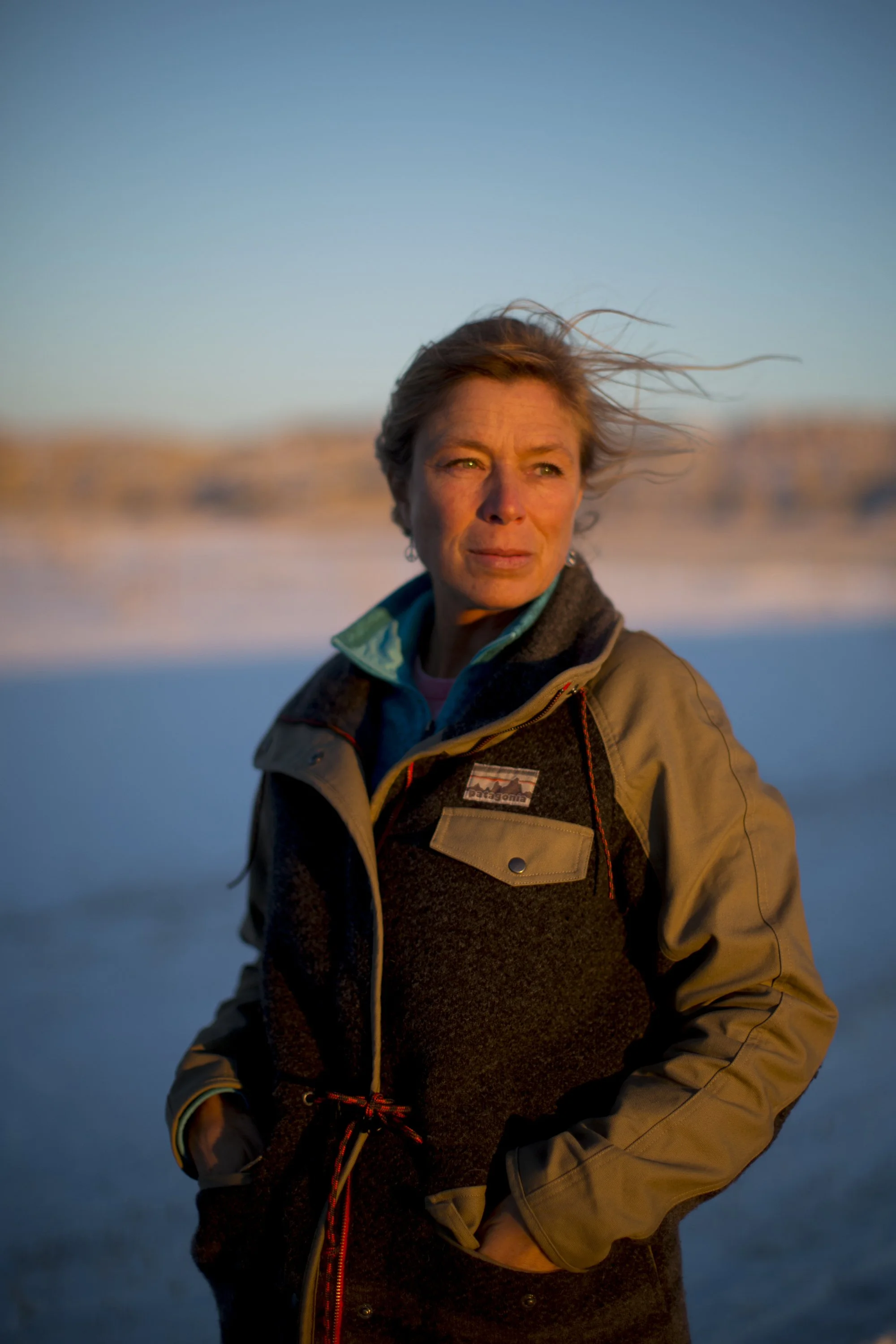 A woman standing outdoors during sunset or sunrise with a blurred landscape in the background, wearing a brown and black outdoor jacket and looking into the distance at the Wild Idea Buffalo Ranch in South Dakota.
