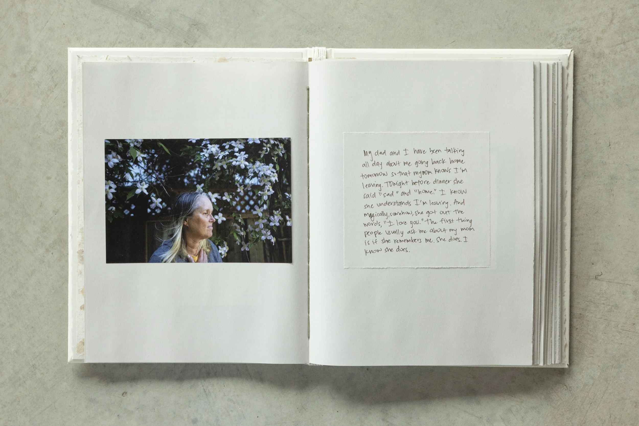 An open scrapbook displaying a printed photo of a woman with gray hair in profile, standing in front of flowering bushes, and a handwritten note on the opposite page.