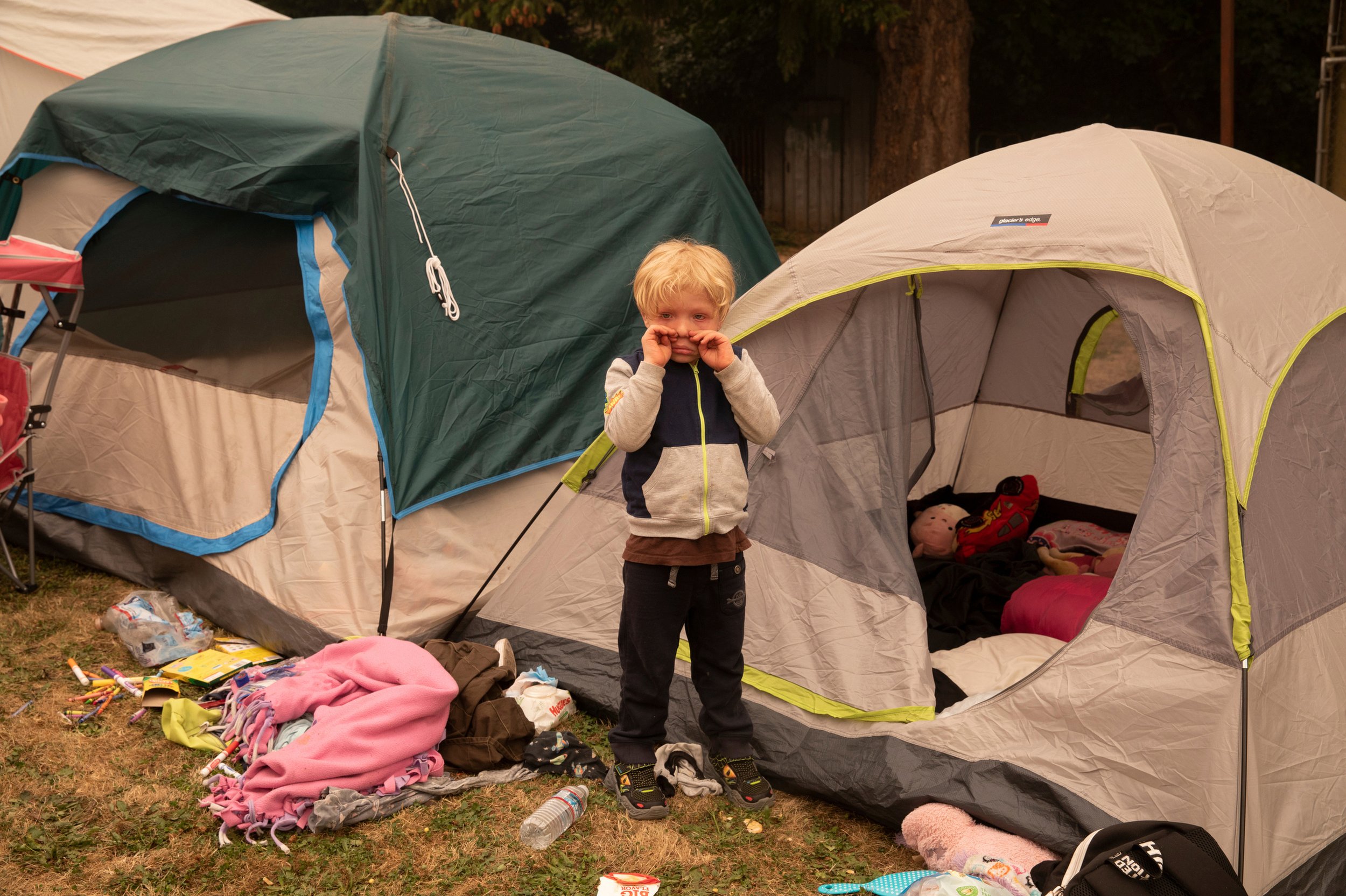 A young boy with blonde hair standing outside among camping tents, with toys and clothes scattered on the ground around him.