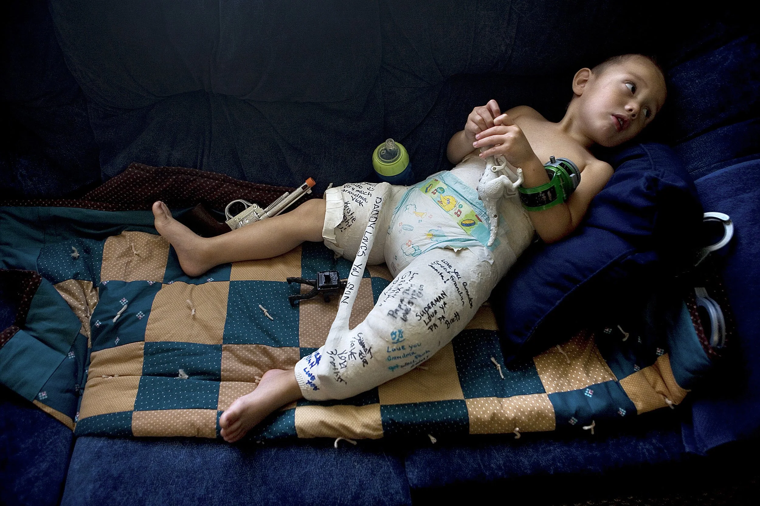 A young boy lying on a quilted blanket on a dark couch. He is shirtless and wearing a diaper with handwritten messages and drawings. The boy has a green smartwatch on his wrist and a blue and green bottle nearby. The blanket and the boy are surrounde