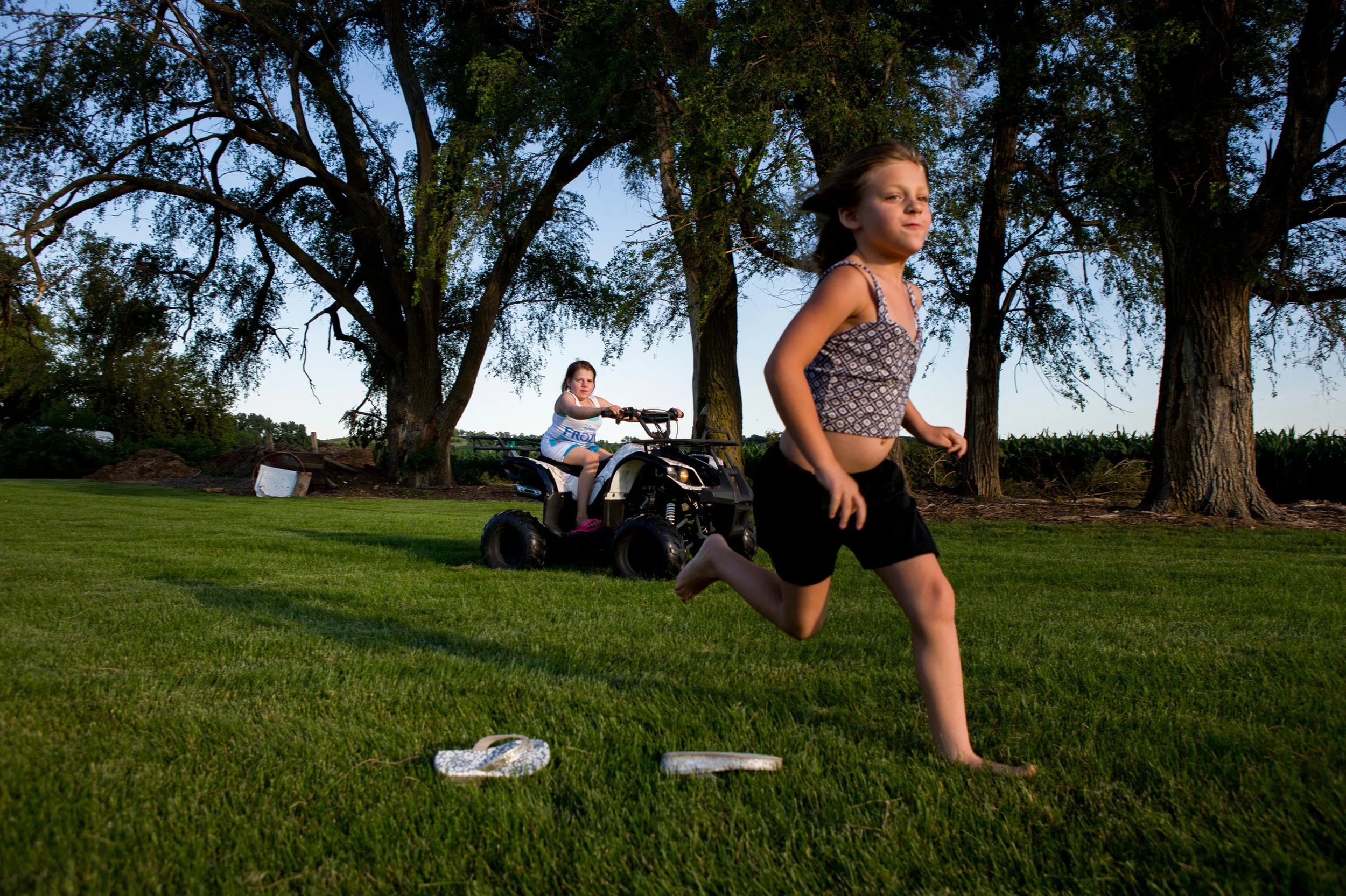 A girl running in the grass while another girl sits on a quad bike behind tall trees during dusk in Nebraska.
