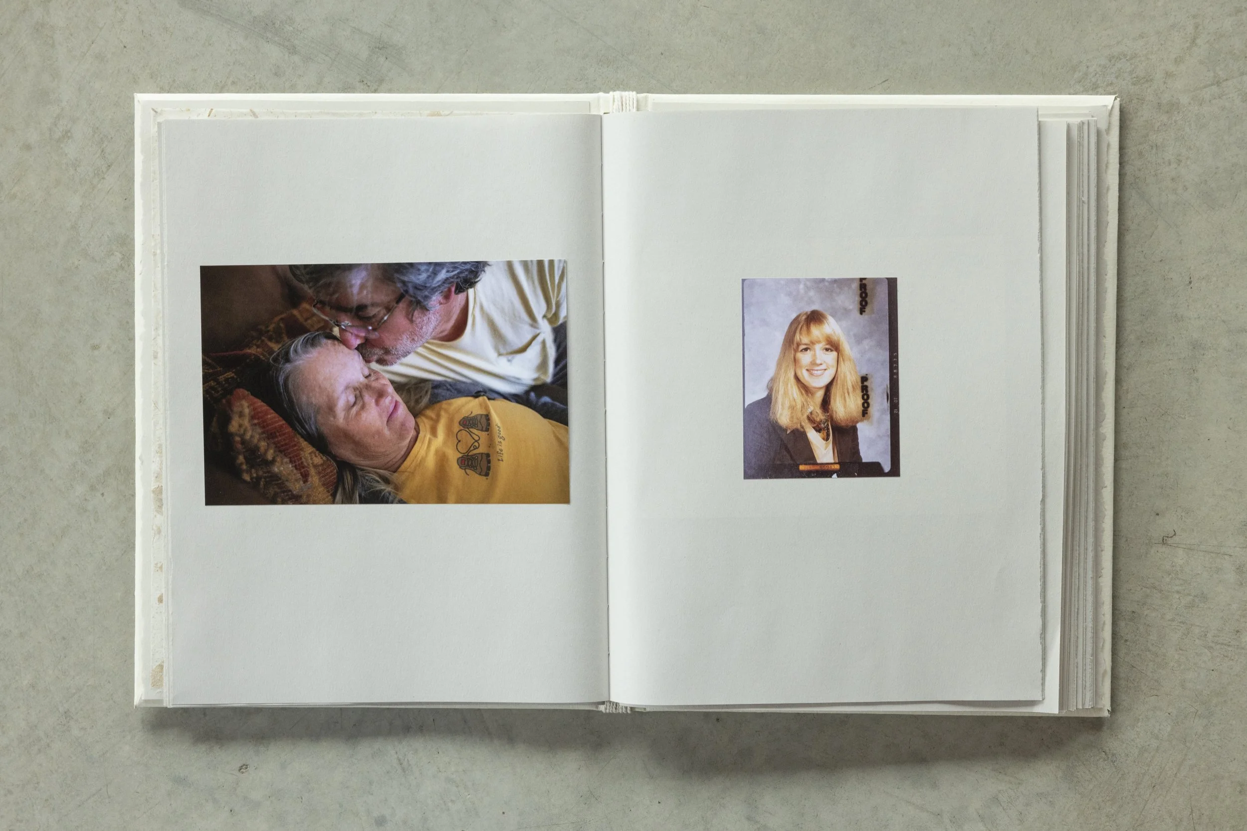 An open photo album with two photographs: one shows a man and woman lying down close to each other with the man kissing the woman's forehead, and the other features a young woman with long red hair and a black blazer smiling at the camera.