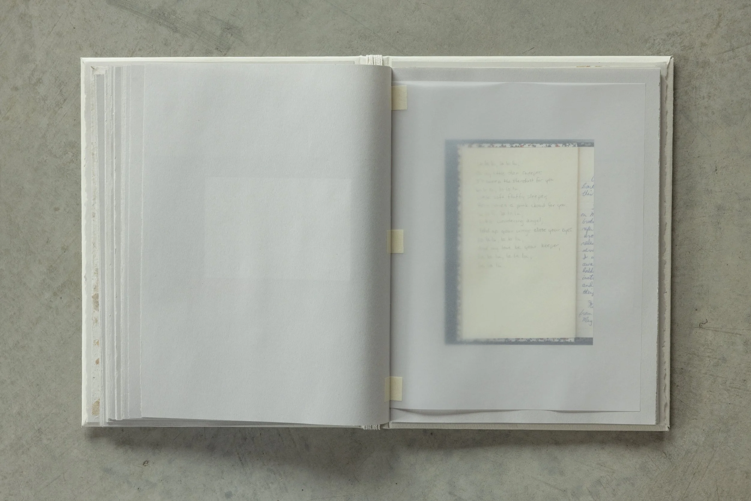 An open white hardcover book resting on a concrete surface, with a blurred photograph and handwritten notes inside.