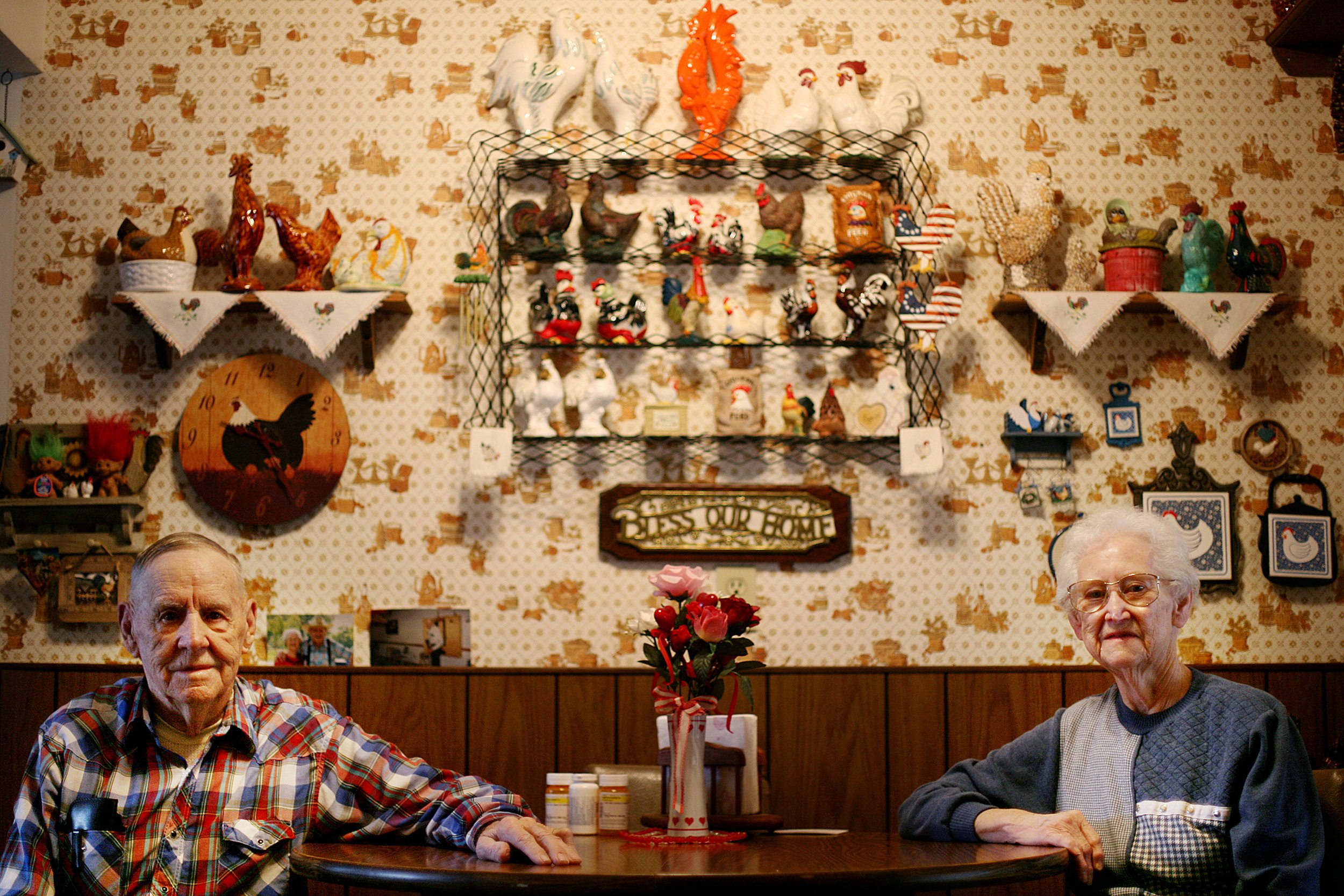 Two elderly people sitting at a table in a room decorated with rooster-themed items, including ceramic roosters on shelves and a rooster clock on the wall. There is a bouquet of pink flowers in the center of the table.