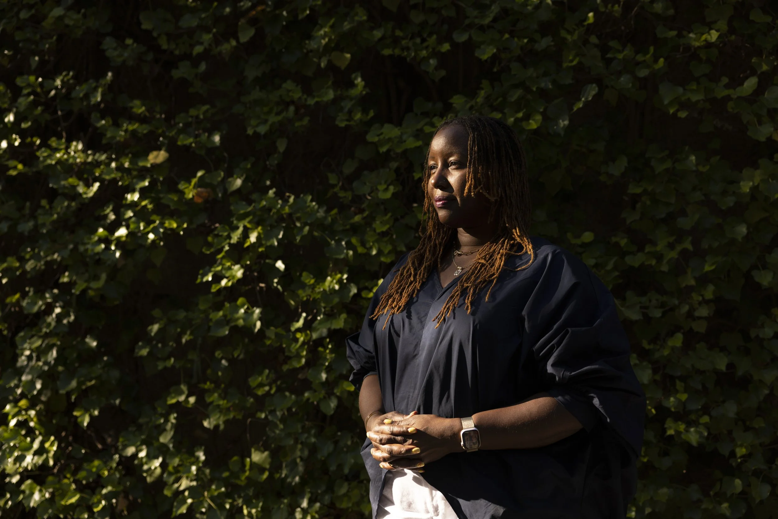 A woman standing outdoors in front of green leafy bushes, looking to the side with sunlight illuminating her face.