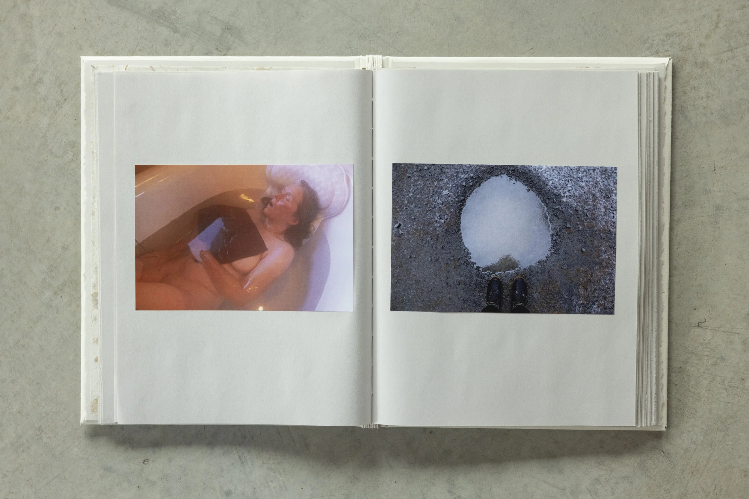 Open photo book with two photographs on white pages; the left shows a woman lying in a bathtub, the right shows feet standing on dirt around a small hole.