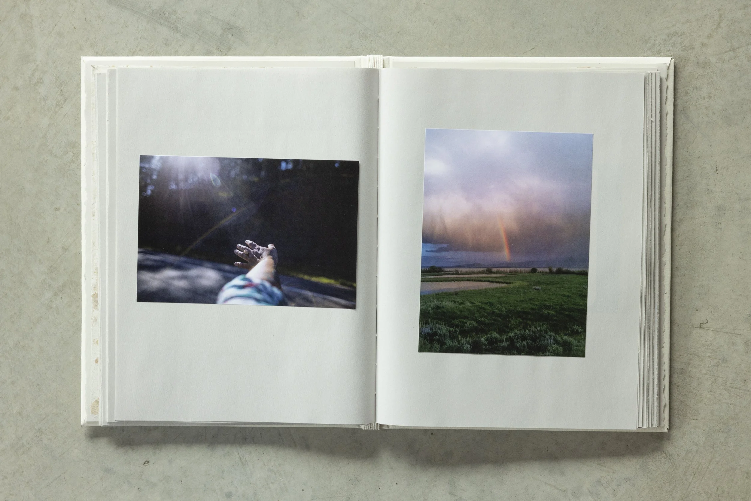 Open photo album showing two nature landscape photographs, one with a rainbow and another with a person's hand reaching towards the sun, on a concrete surface.
