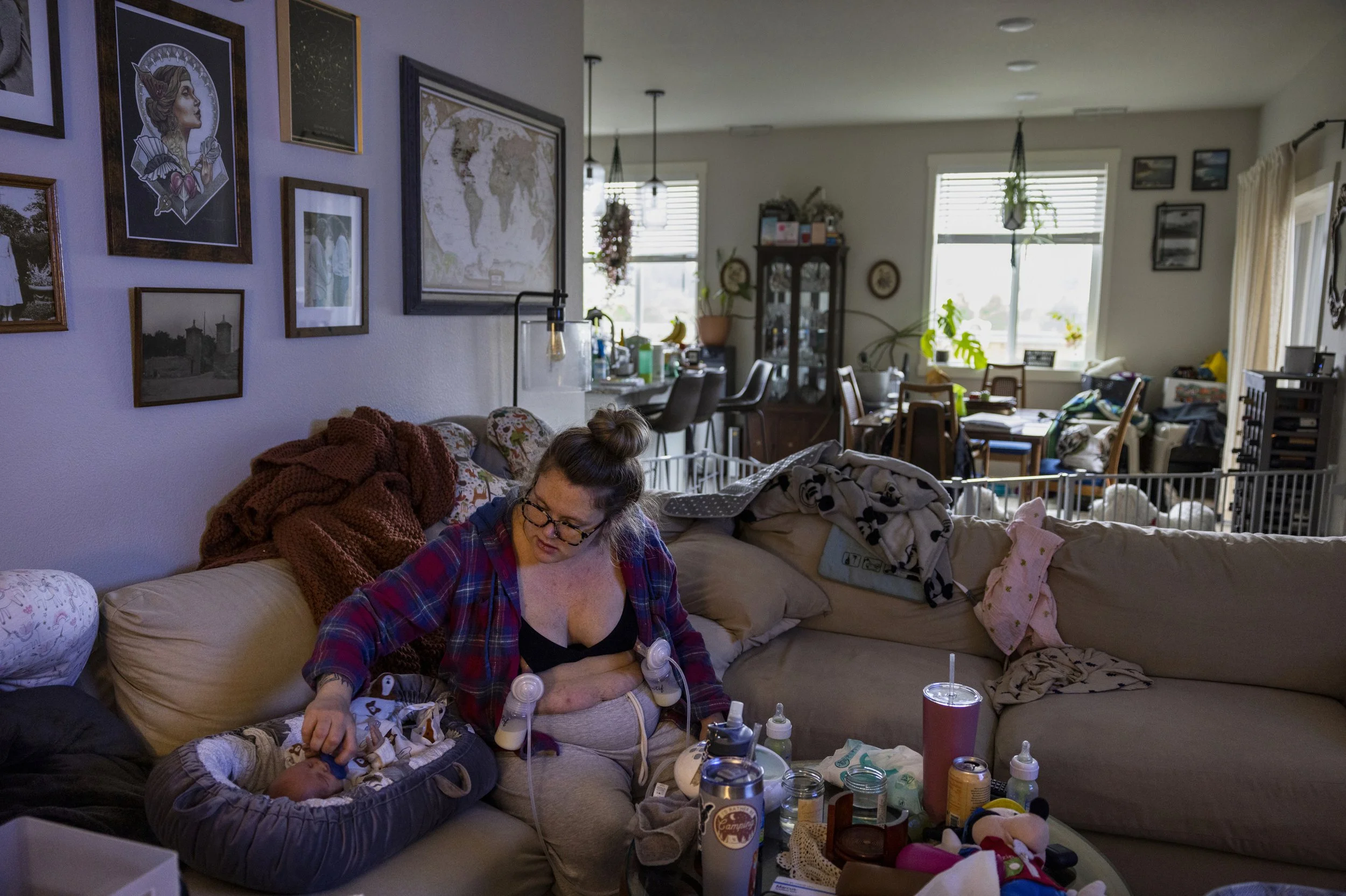 A woman sitting on a beige couch with a baby, holding feeding bottles, in a cluttered living room with art on the walls and scattered objects in Oregon.