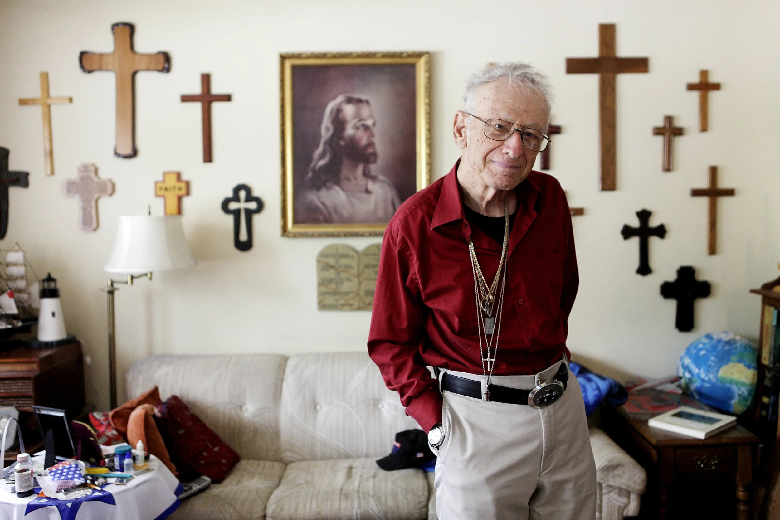 An elderly man with glasses and white hair standing in a living room. Behind him on the wall are multiple religious crosses and a framed photo of Jesus Christ. The room has a beige sofa, a side table with a lamp, and clutter on a table and chair.