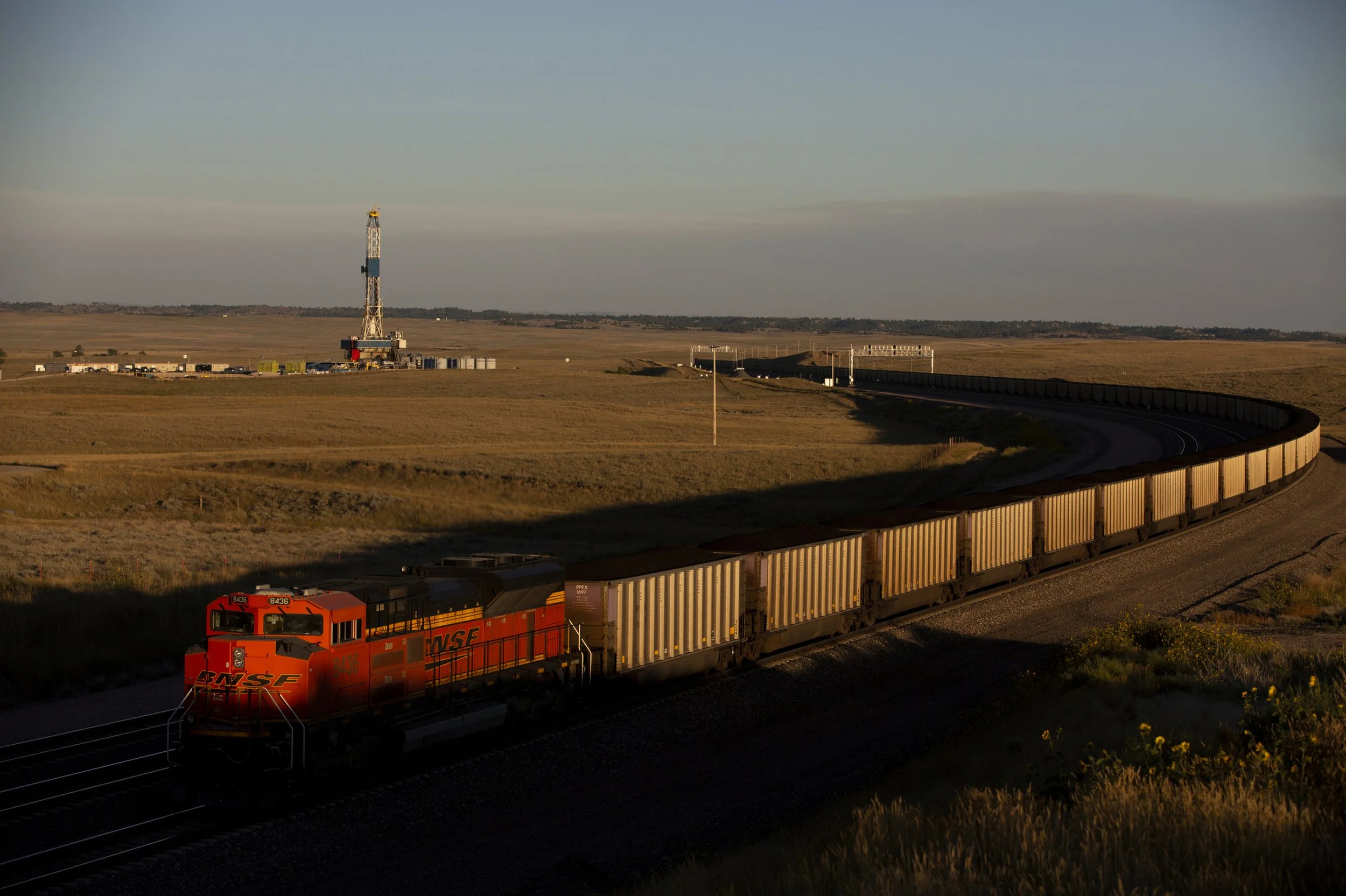A red and black BNSF freight train moving through a rural landscape with a long curved track, a tall radio tower, power lines, and a wide open sky during sunset in Wyoming. 
